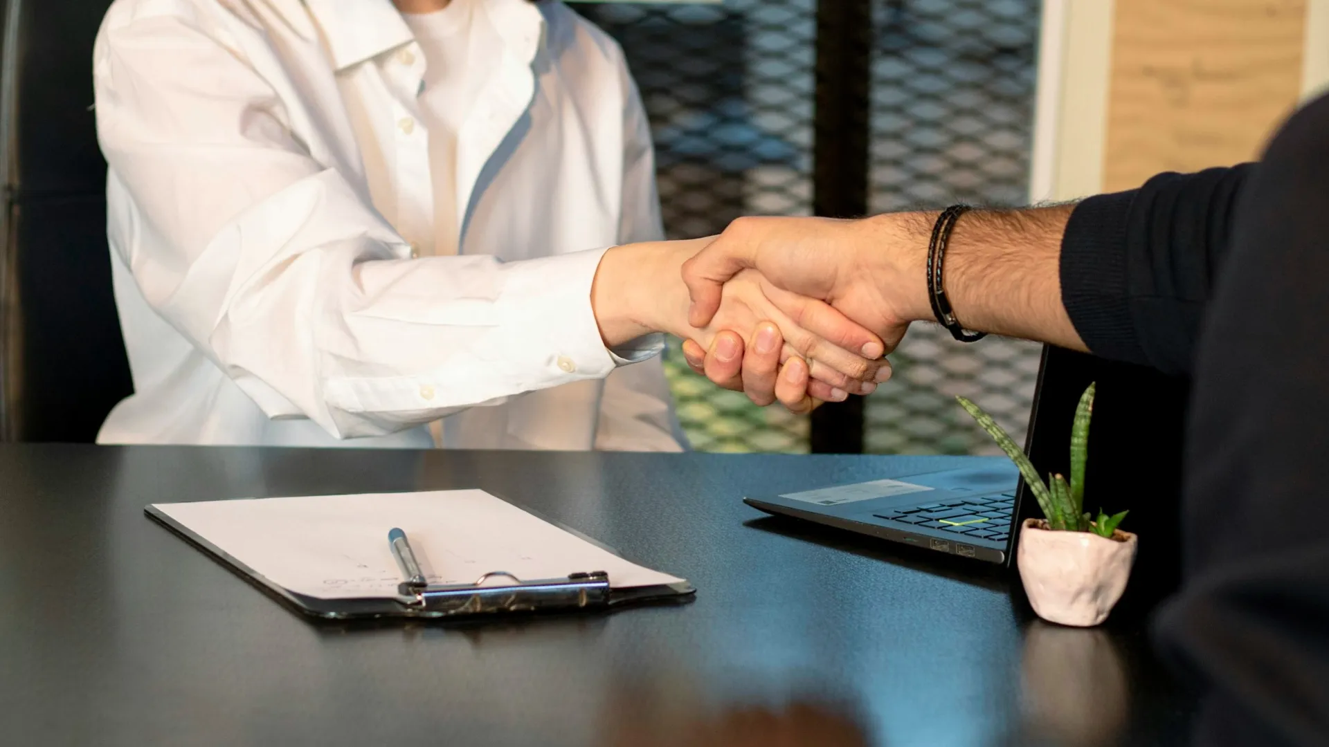 Two professionals shaking hands over a desk with laptop, clipboard, and small plant in an office setting.