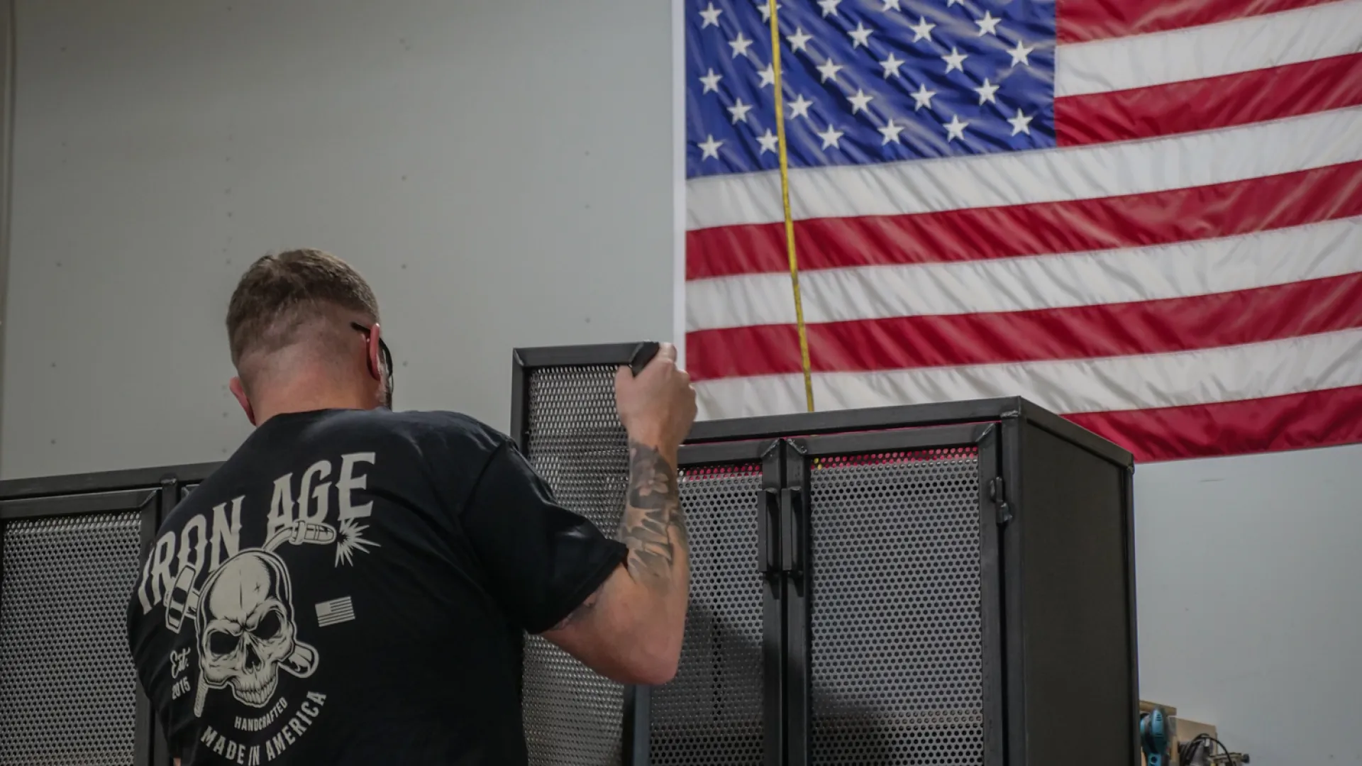 Worker installing metal cabinet doors in workshop with American flag on wall in the background.