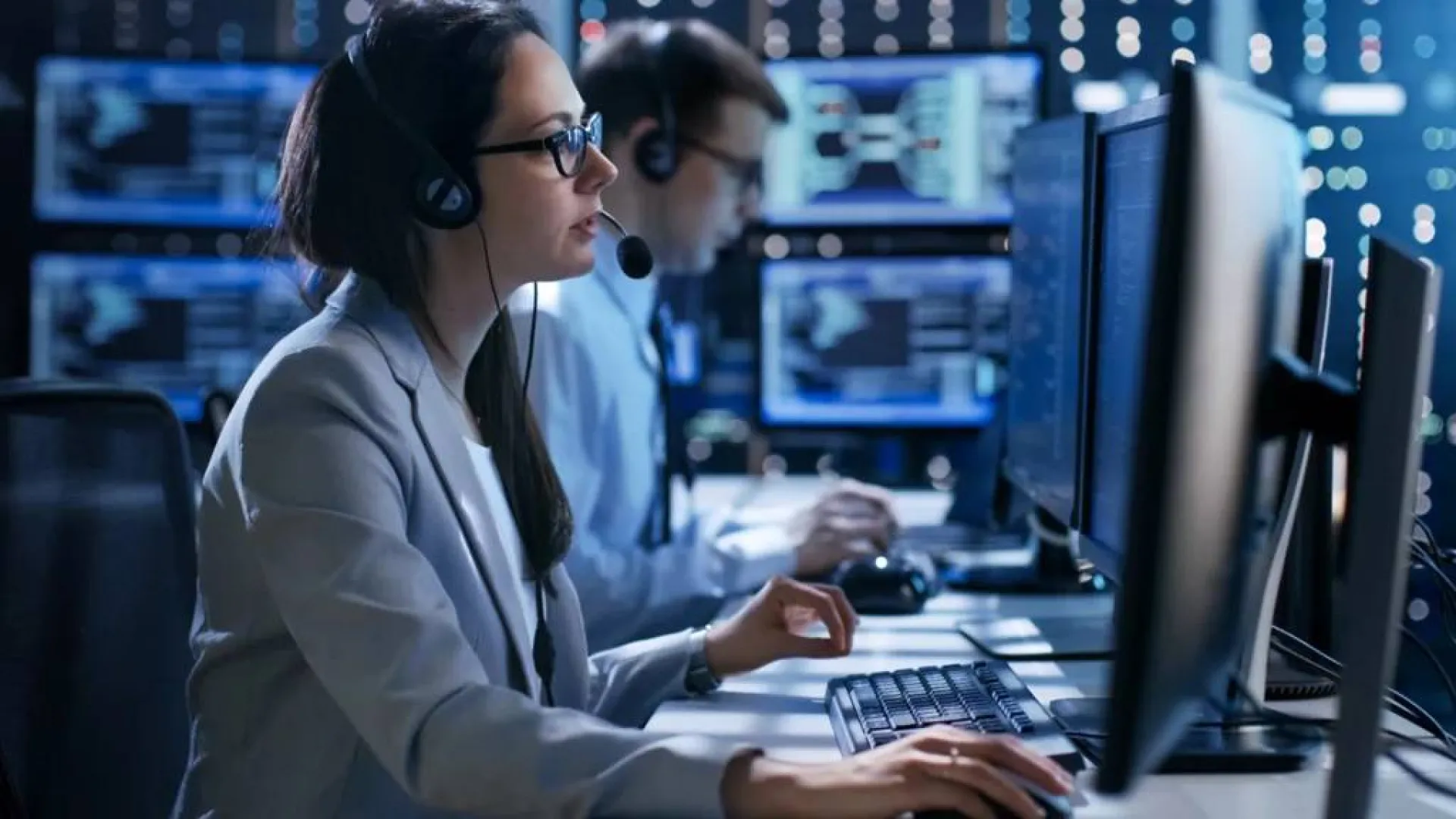 Woman and man in headset working on multiple computers in a modern data center or security operations room.