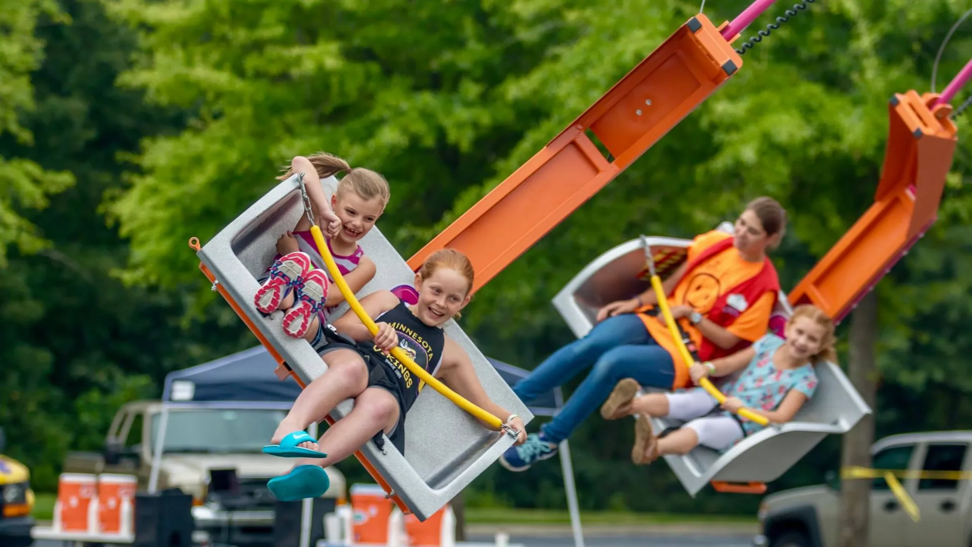 Children enjoy a swinging amusement park ride on a bright day with green trees in the background.