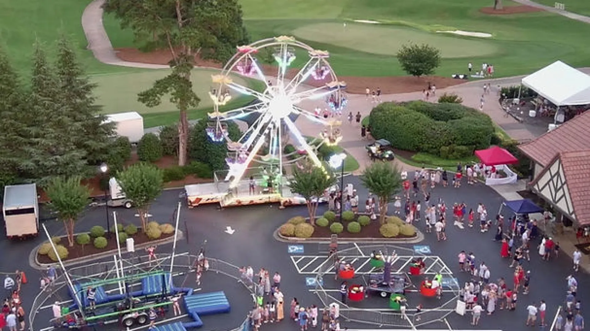 Evening view of a small carnival with illuminated Ferris wheels and rides near a golf course with green fairways.