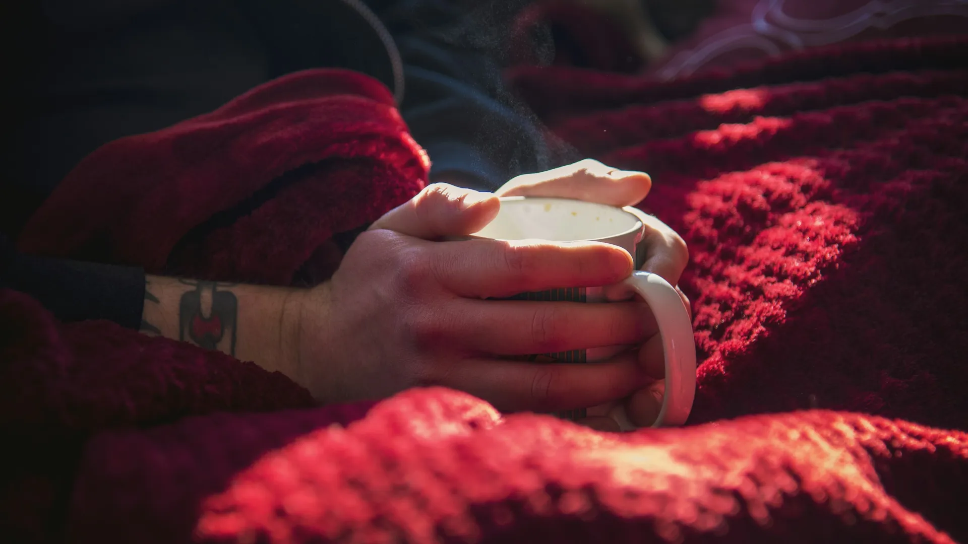 Person holding a steaming cup wrapped in a cozy red blanket with a tattooed wrist visible in soft light.
