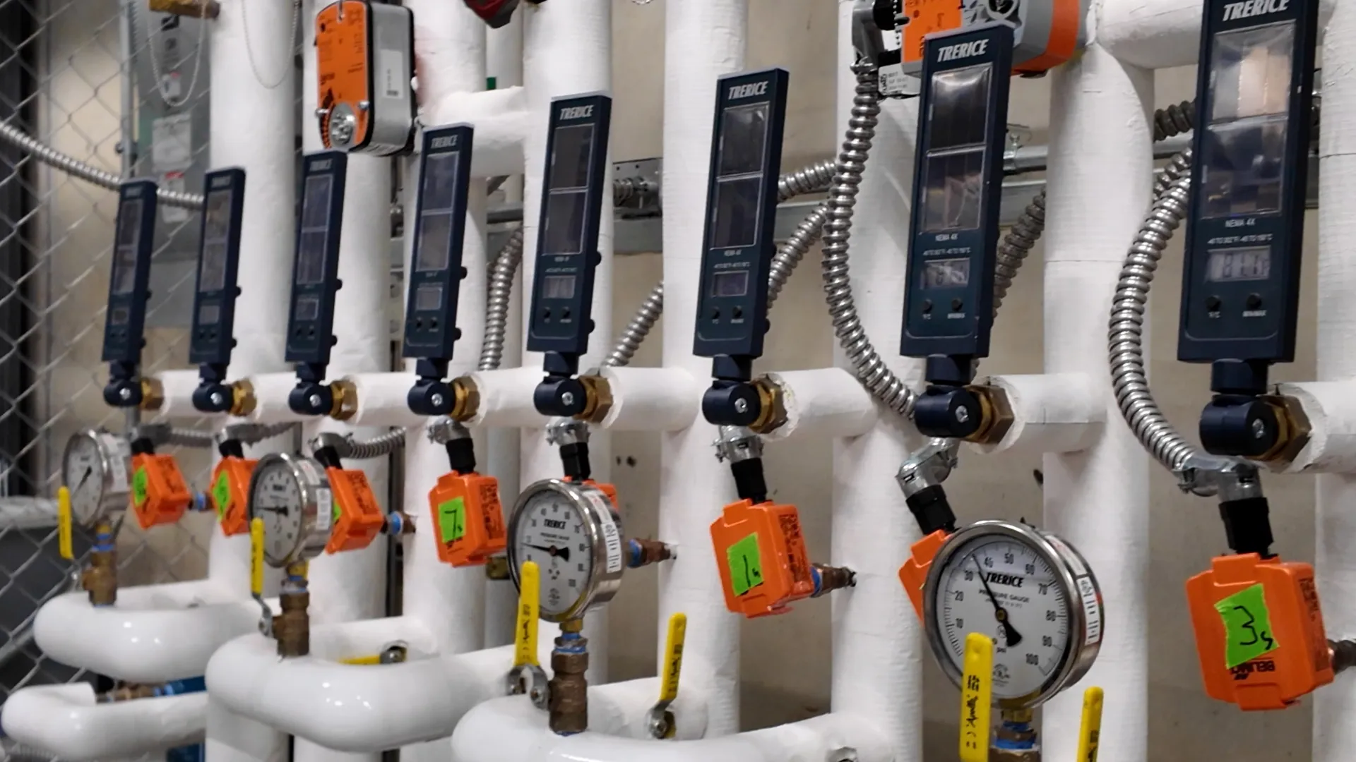 Row of pressure gauges and digital controllers on white industrial piping with metal conduit in a mechanical room.