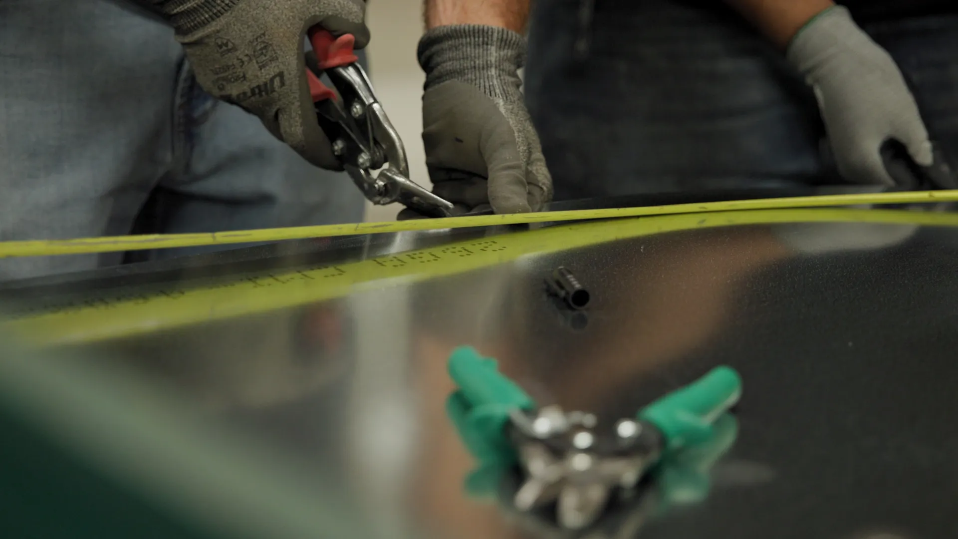 Workers wearing gloves using pliers to bend metal sheet with measuring tape on table.