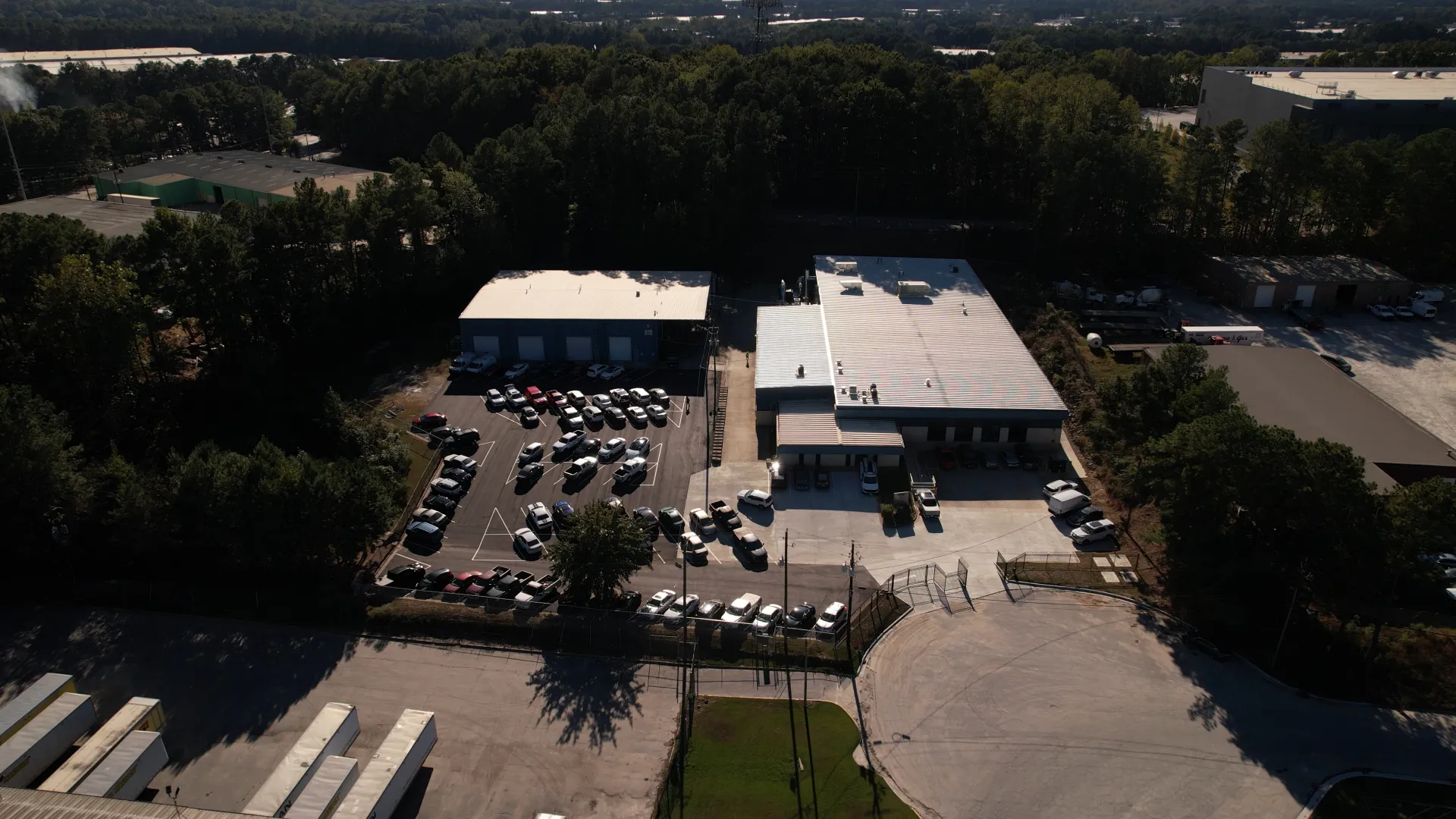 Aerial view of an industrial facility with warehouse buildings, parked cars, surrounded by trees and roads.