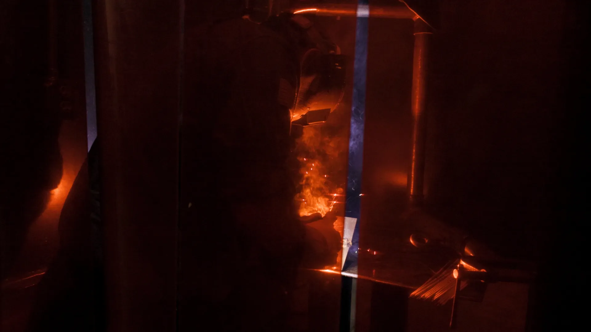 Person welding metal in a dark workshop with bright orange sparks and protective gear