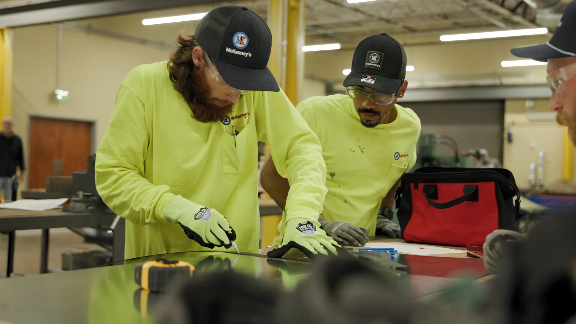 Two men in yellow shirts and gloves working on a metal sheet in an industrial workshop setting.