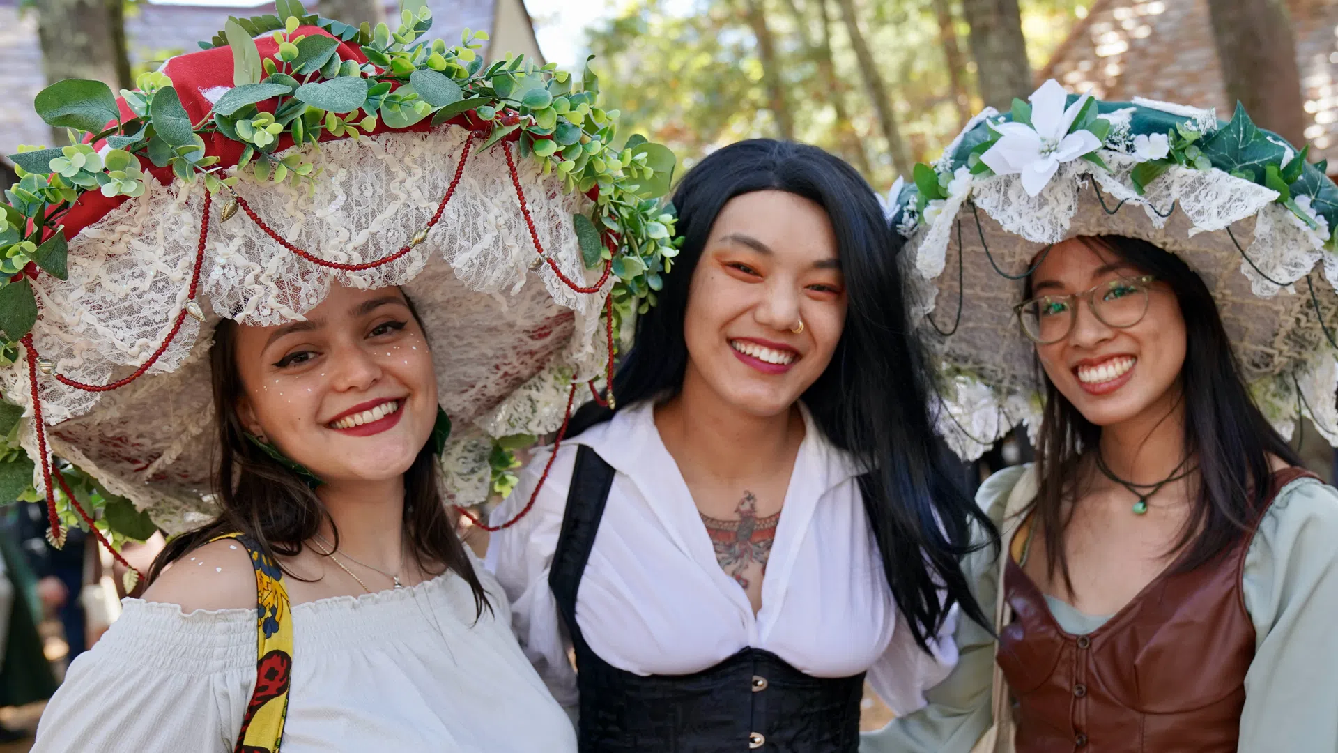 Three smiling women wearing large floral hats and Renaissance-style clothing outdoors in a wooded area