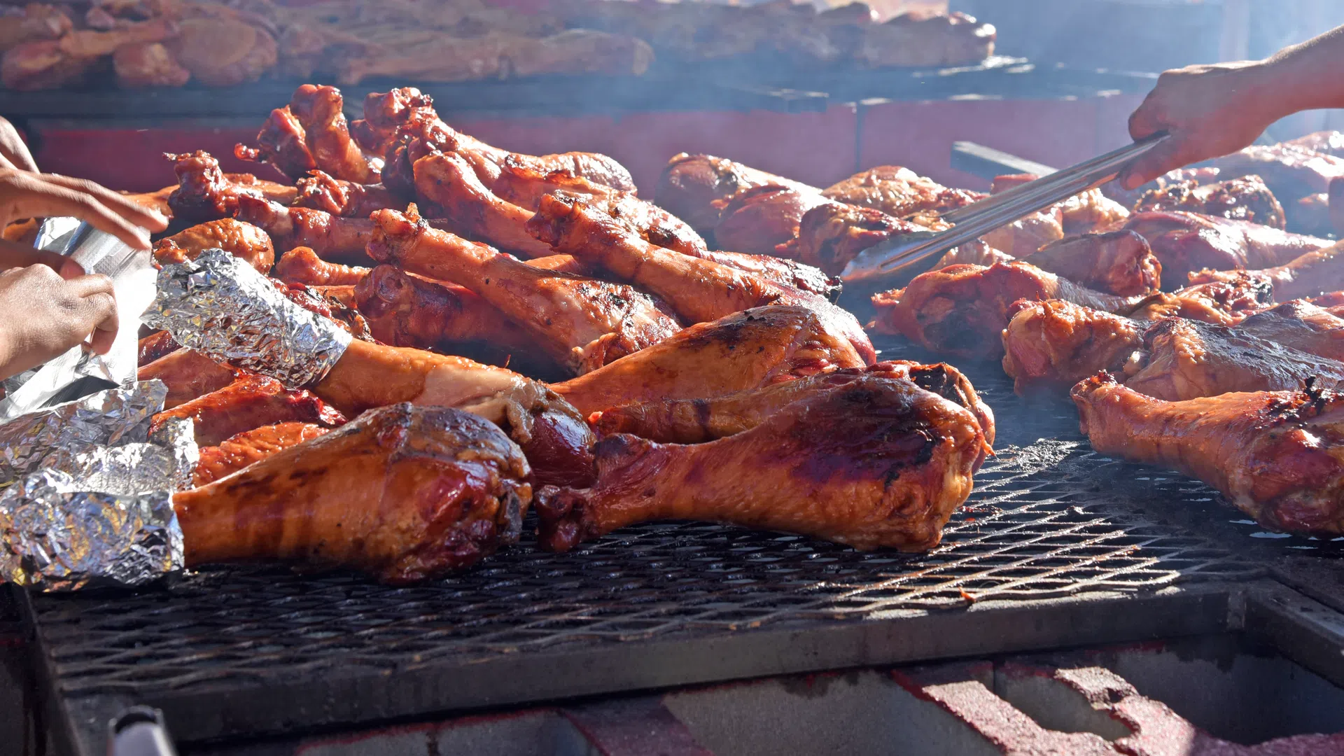 Grilled turkey legs cooking over a charcoal barbecue with hands turning meat using tongs on an outdoor grill.