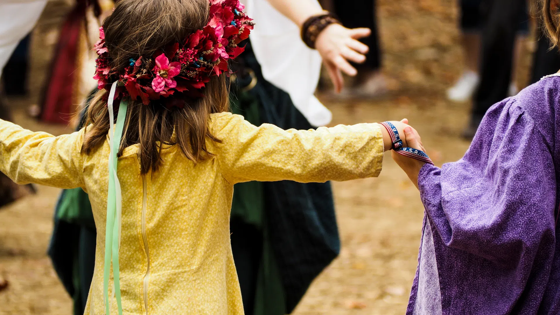 Children in colorful medieval costumes holding hands and dancing outdoors during a festive event.