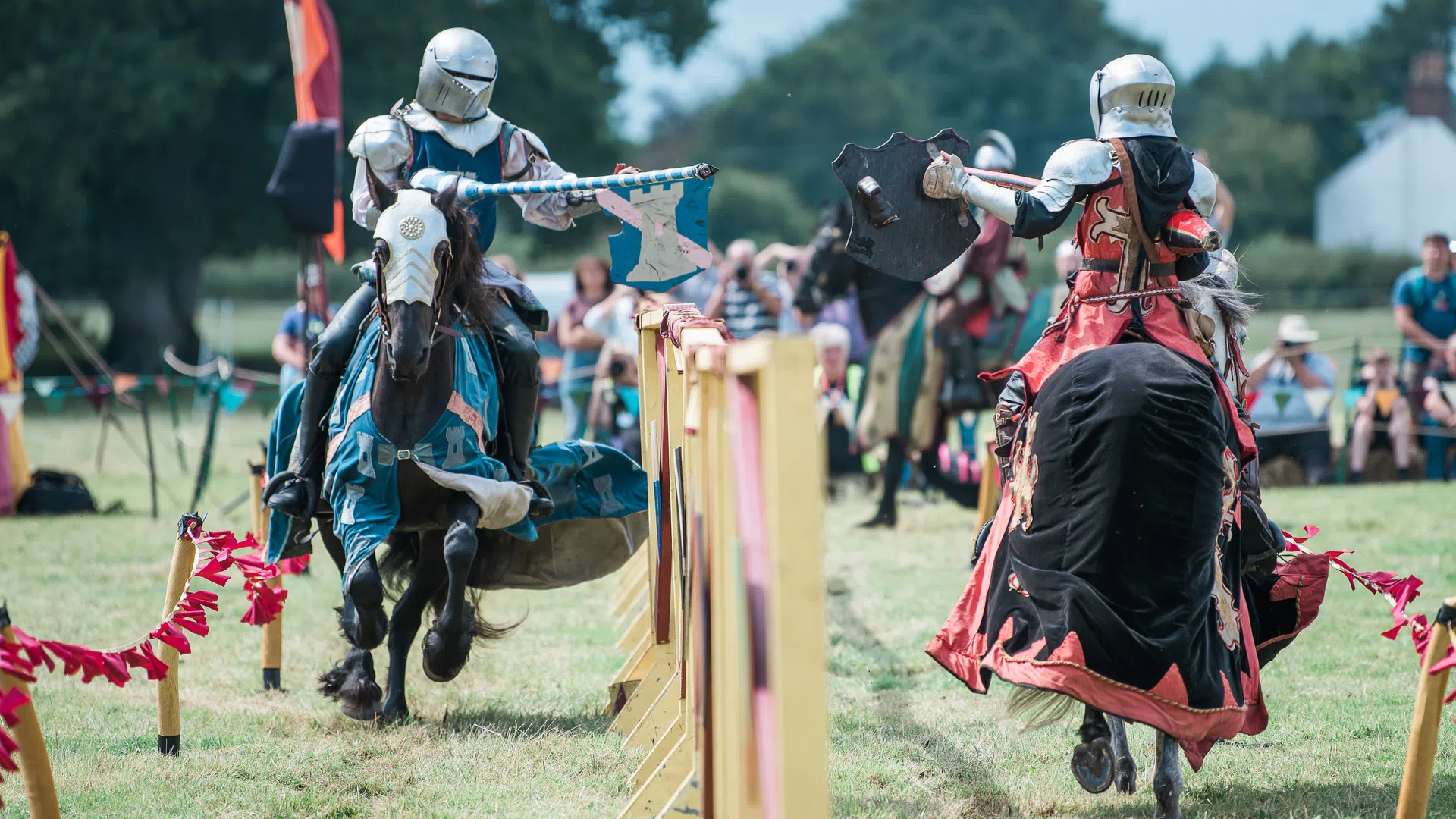 Two knights in armor jousting on horseback during a medieval reenactment in a grassy field with spectators.
