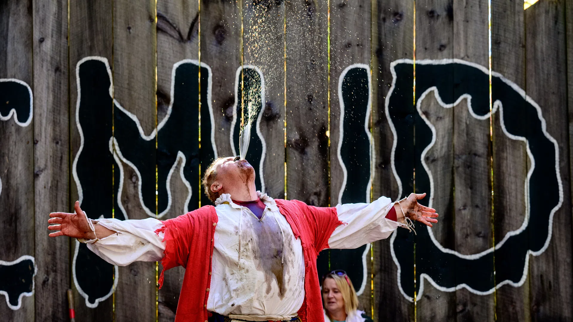 Performer in red and white costume with arms outstretched and water sprayed from mouth in front of wooden fence with bold graffiti text.