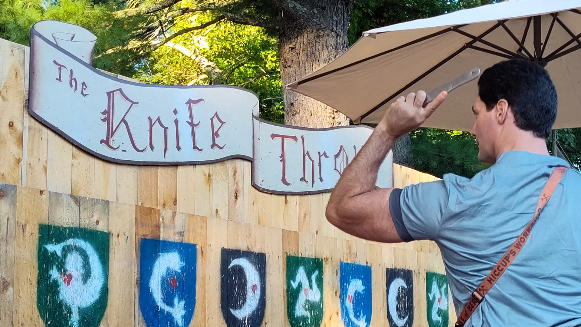 Man aiming a knife at a wooden target board labeled The Knife Throw with shield symbols outdoors under an umbrella.