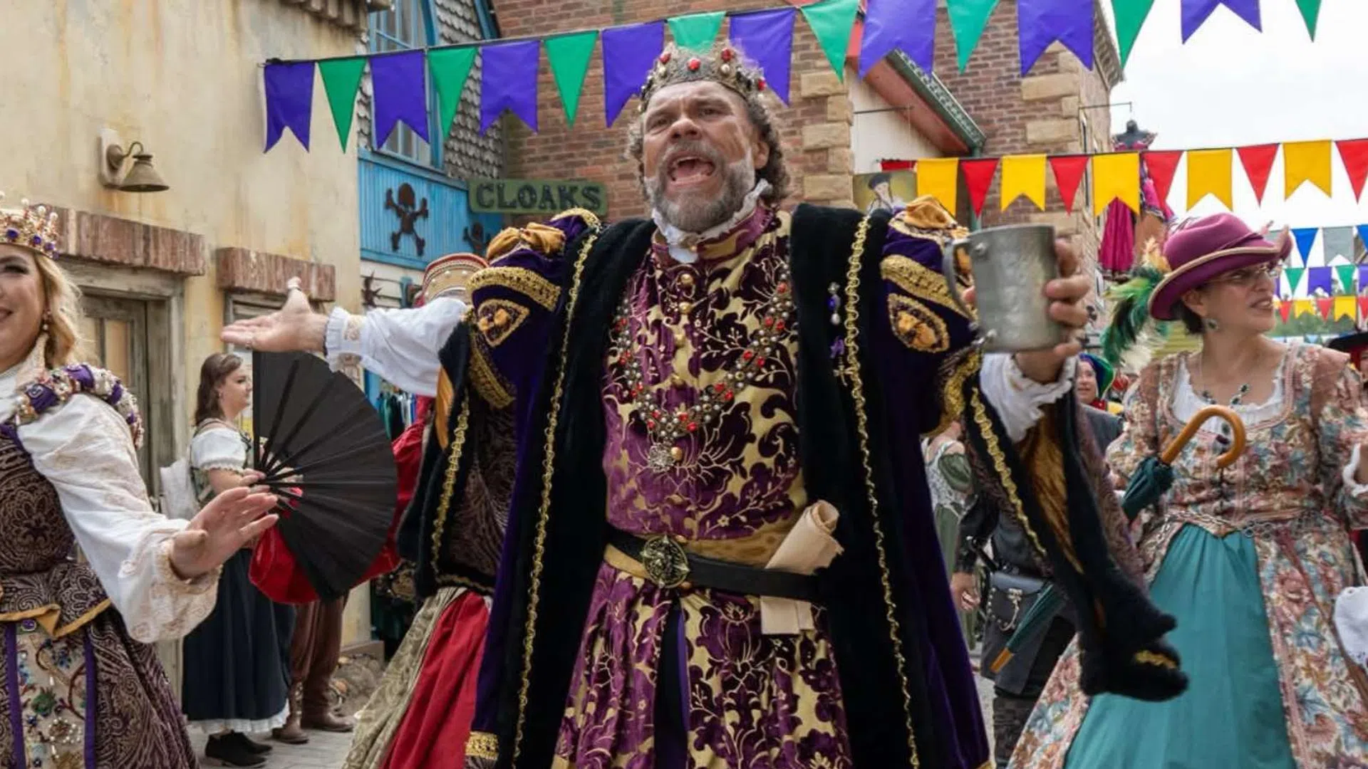 People in colorful Renaissance costumes perform joyfully at a lively street festival with vibrant banners overhead.