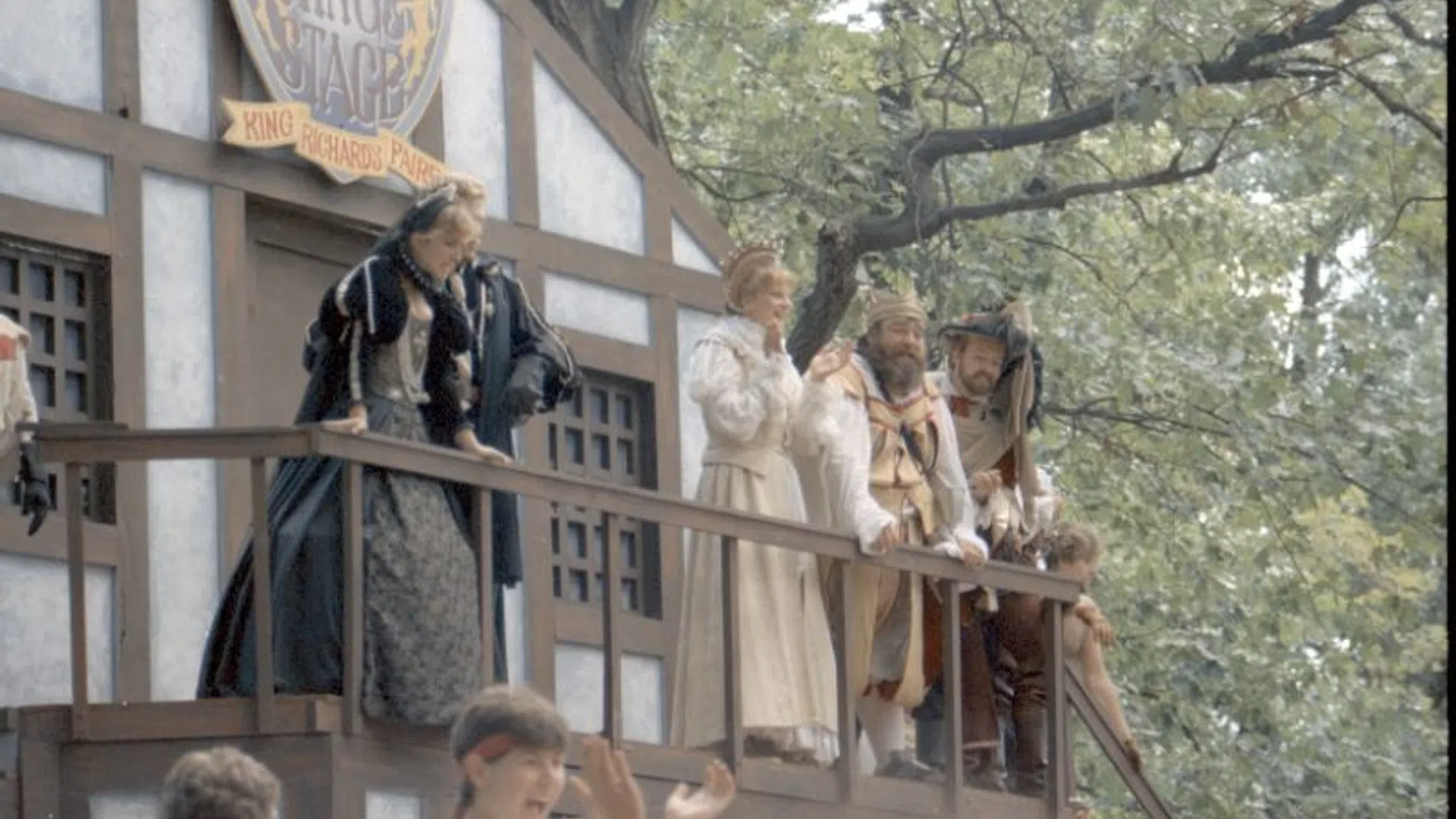 Actors in Renaissance costumes perform on a wooden stage at King Richard's Faire surrounded by green trees.