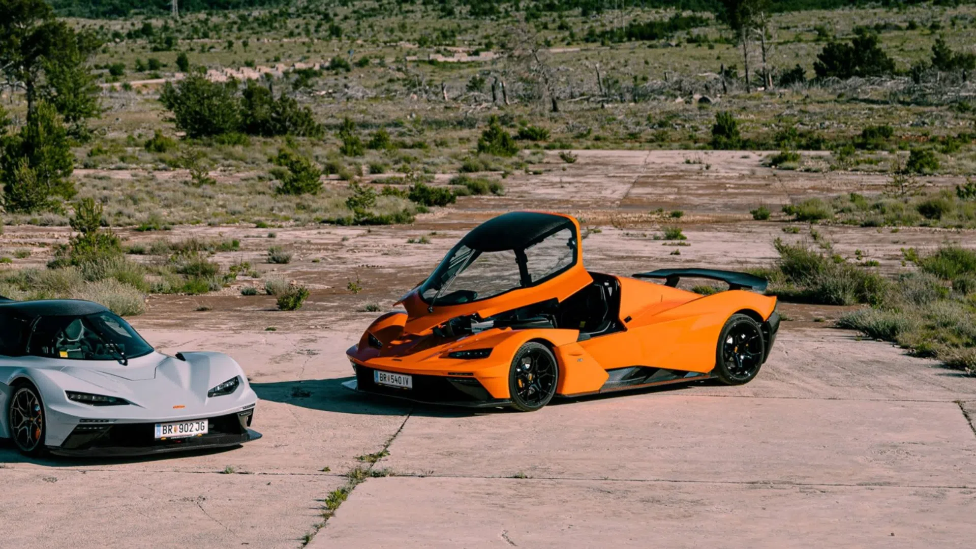 Two sleek sports cars, one gray and one orange with an open roof, parked on concrete in a mountainous area with wind turbines.
