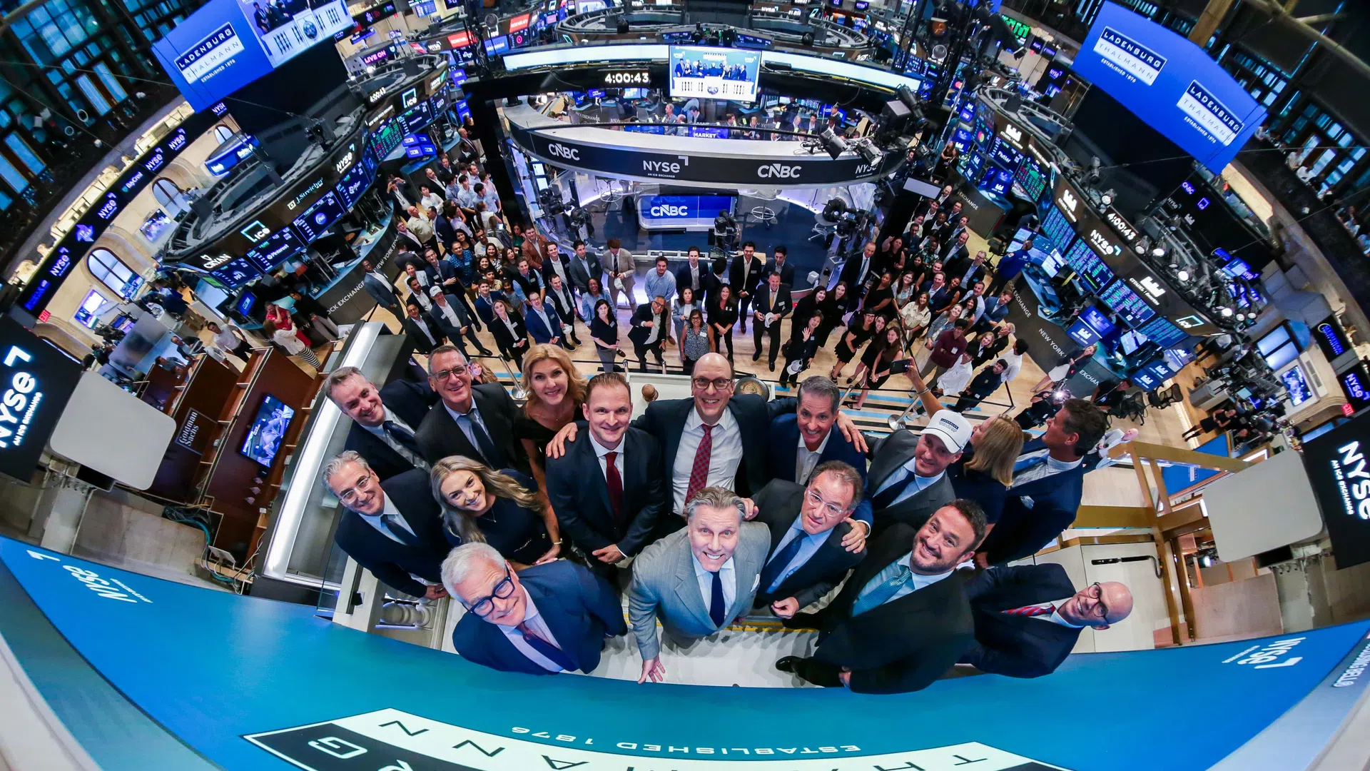 Group of business professionals gathered at the New York Stock Exchange trading floor with digital screens and NYSE branding.