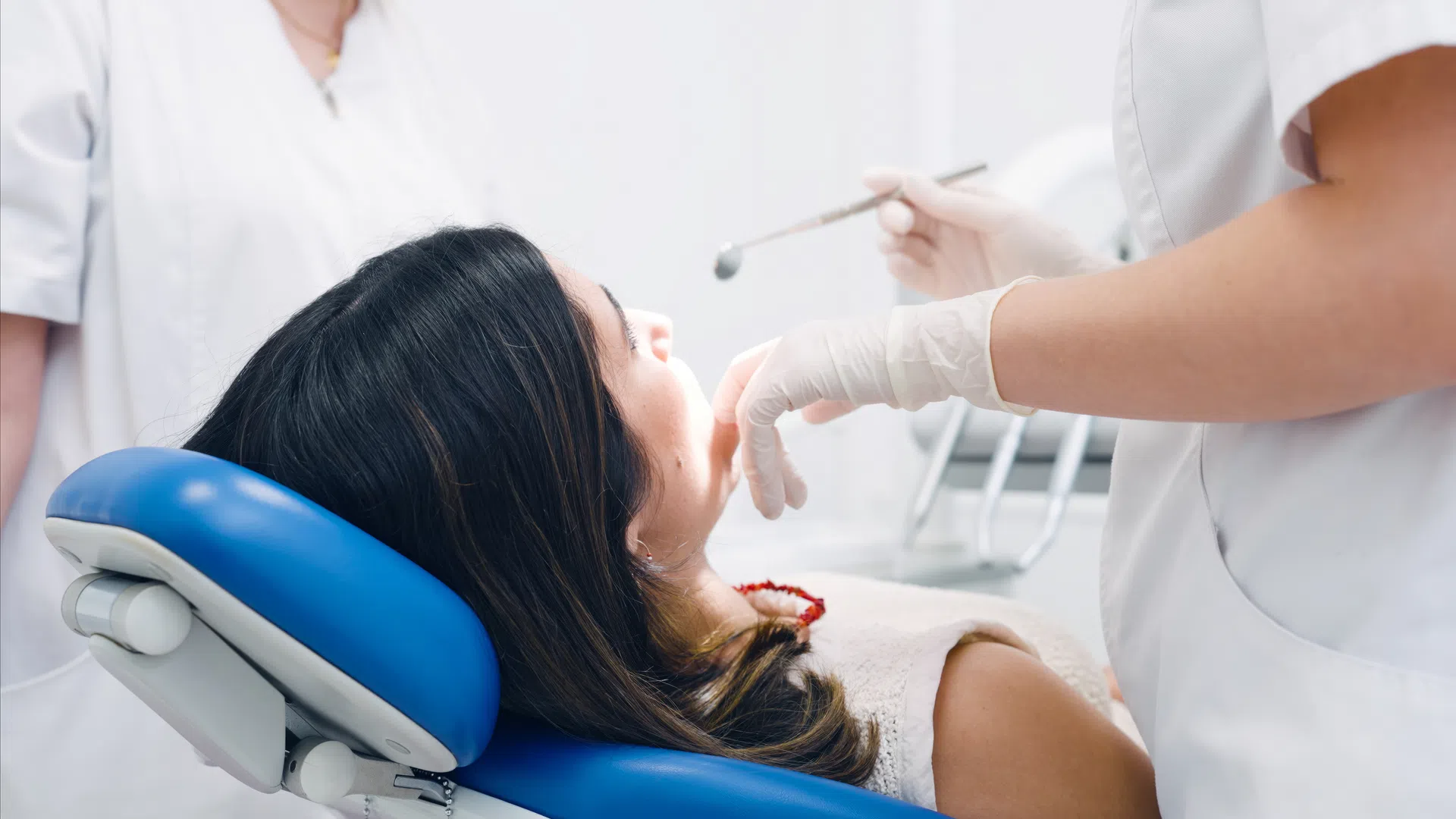 Female patient receiving dental examination from dentist wearing gloves in clinic with blue chair.