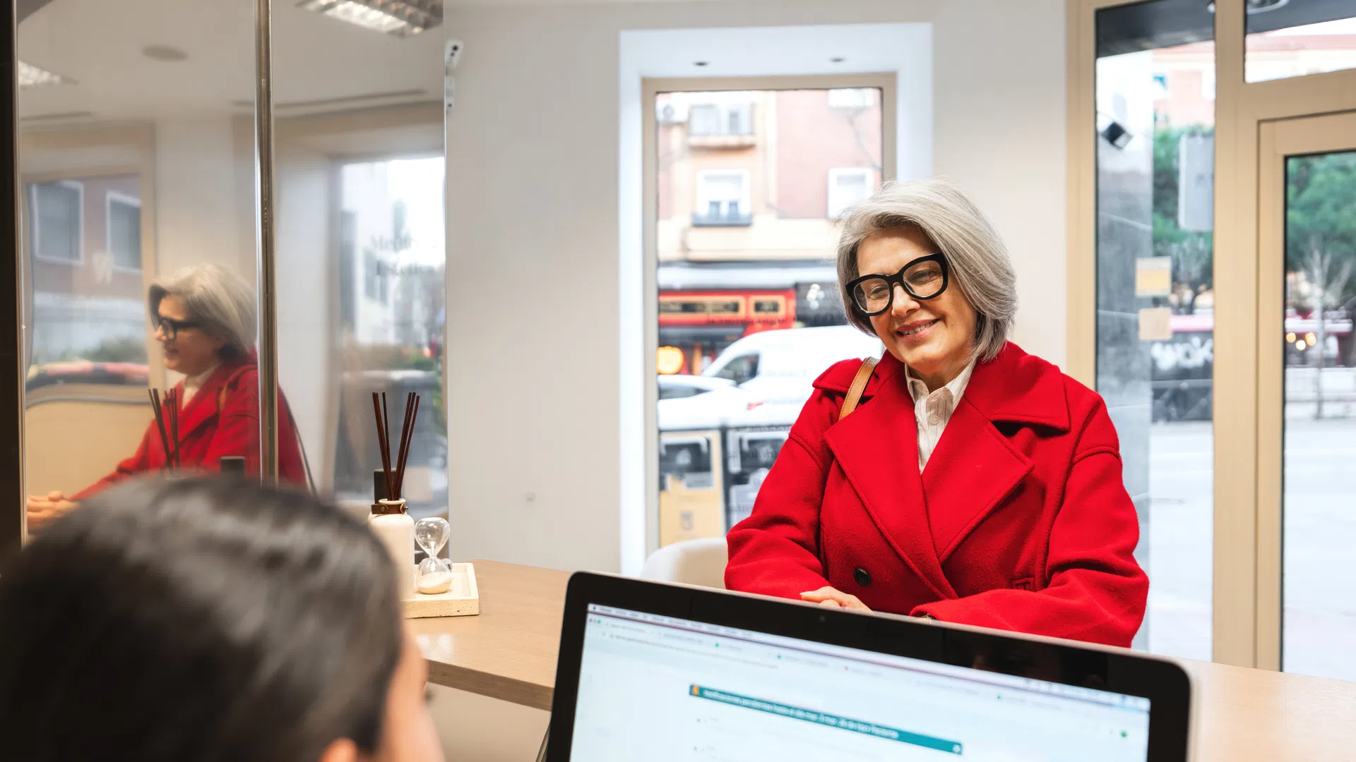 Senior woman in red coat and glasses smiling during consultation at office reception desk.