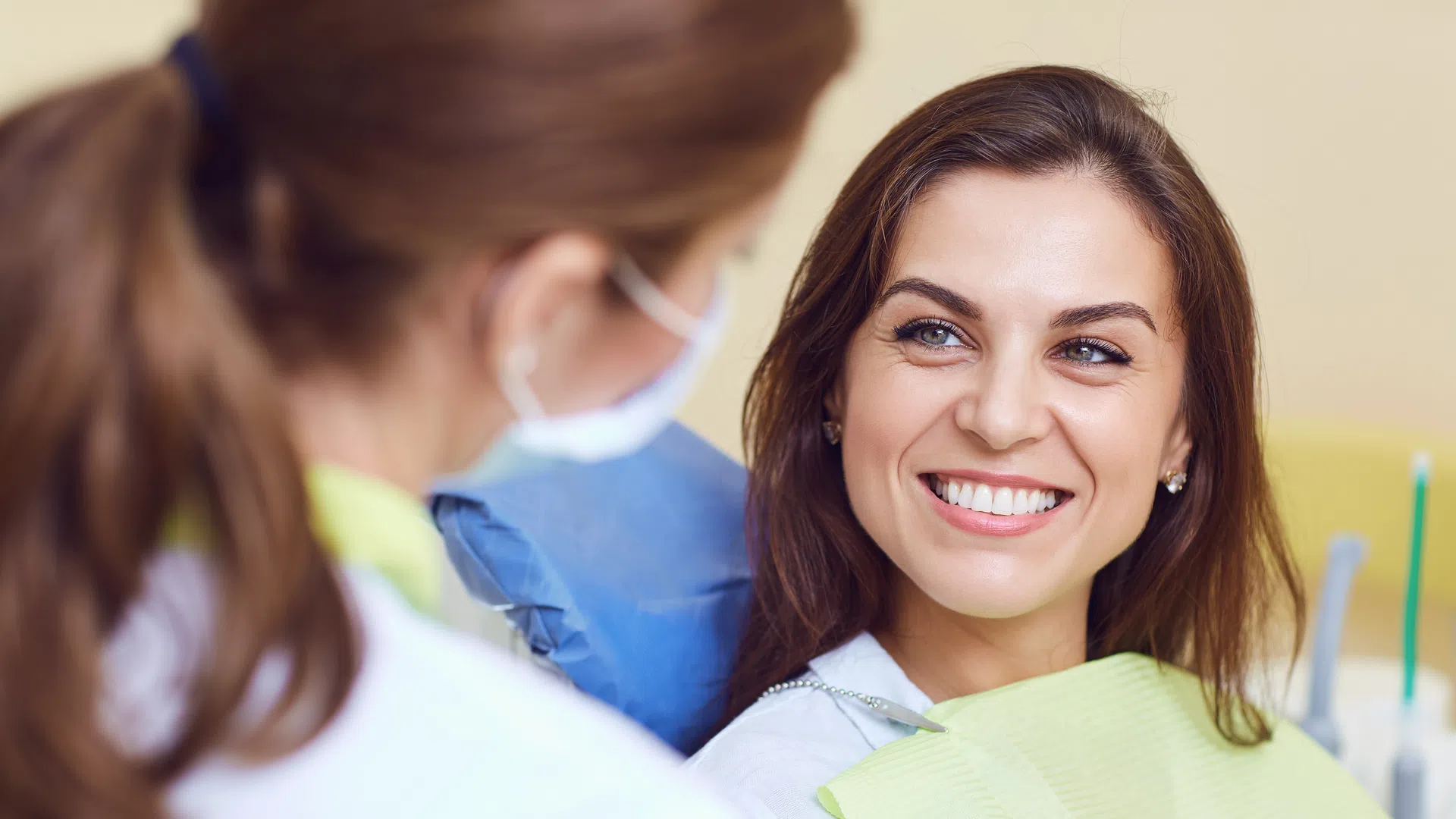 Smiling woman in dental chair with bib talks to female dentist wearing mask in clinic