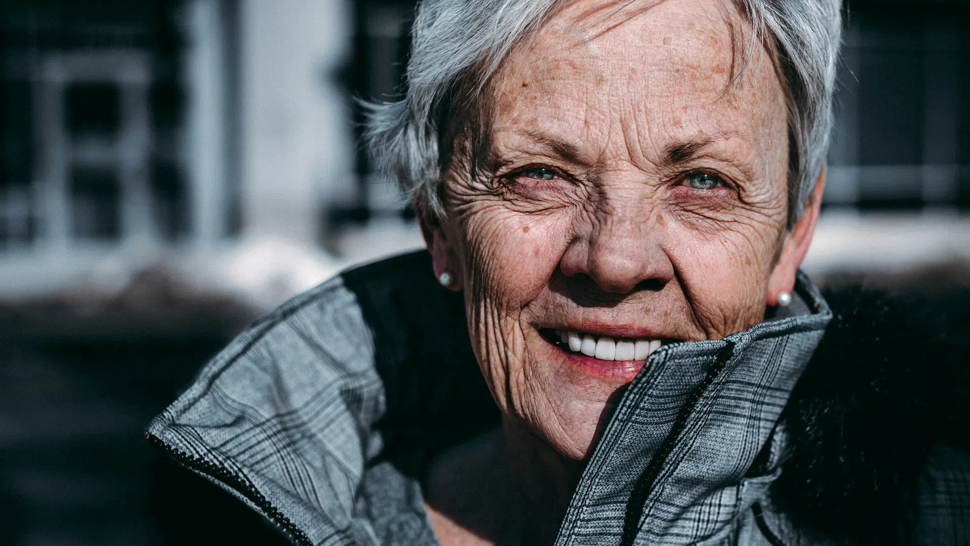 Close-up portrait of a smiling elderly woman with short gray hair wearing a gray jacket outdoors.