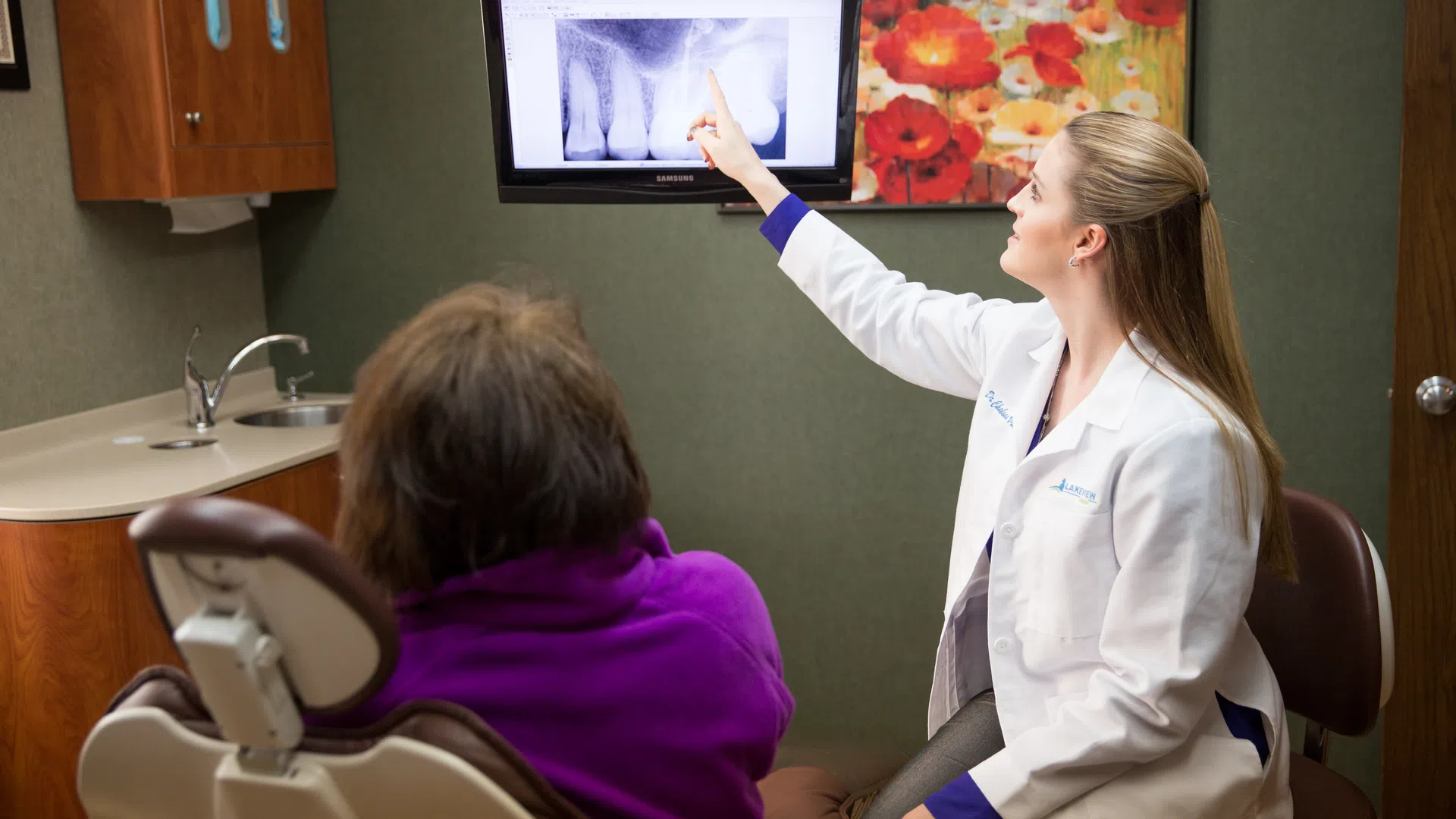 Dentist explains dental X-ray results to patient in office with floral painting and wooden cabinets.