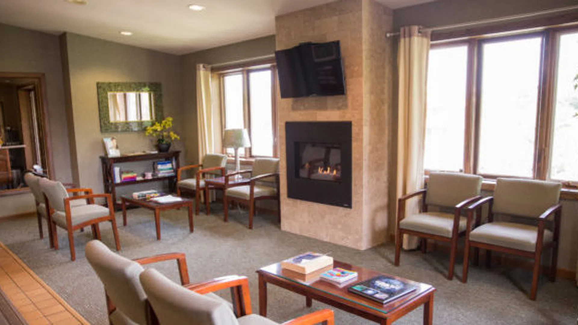 Cozy waiting room with cushioned chairs, a fireplace, flat-screen TV, and natural light from large windows.