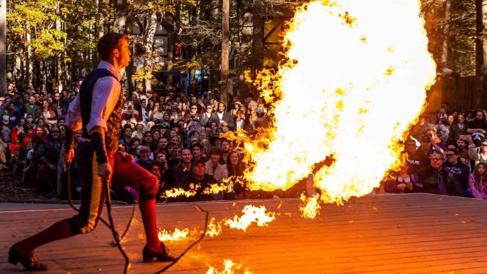Performer breathing fire on stage in front of a large outdoor audience during daytime event.