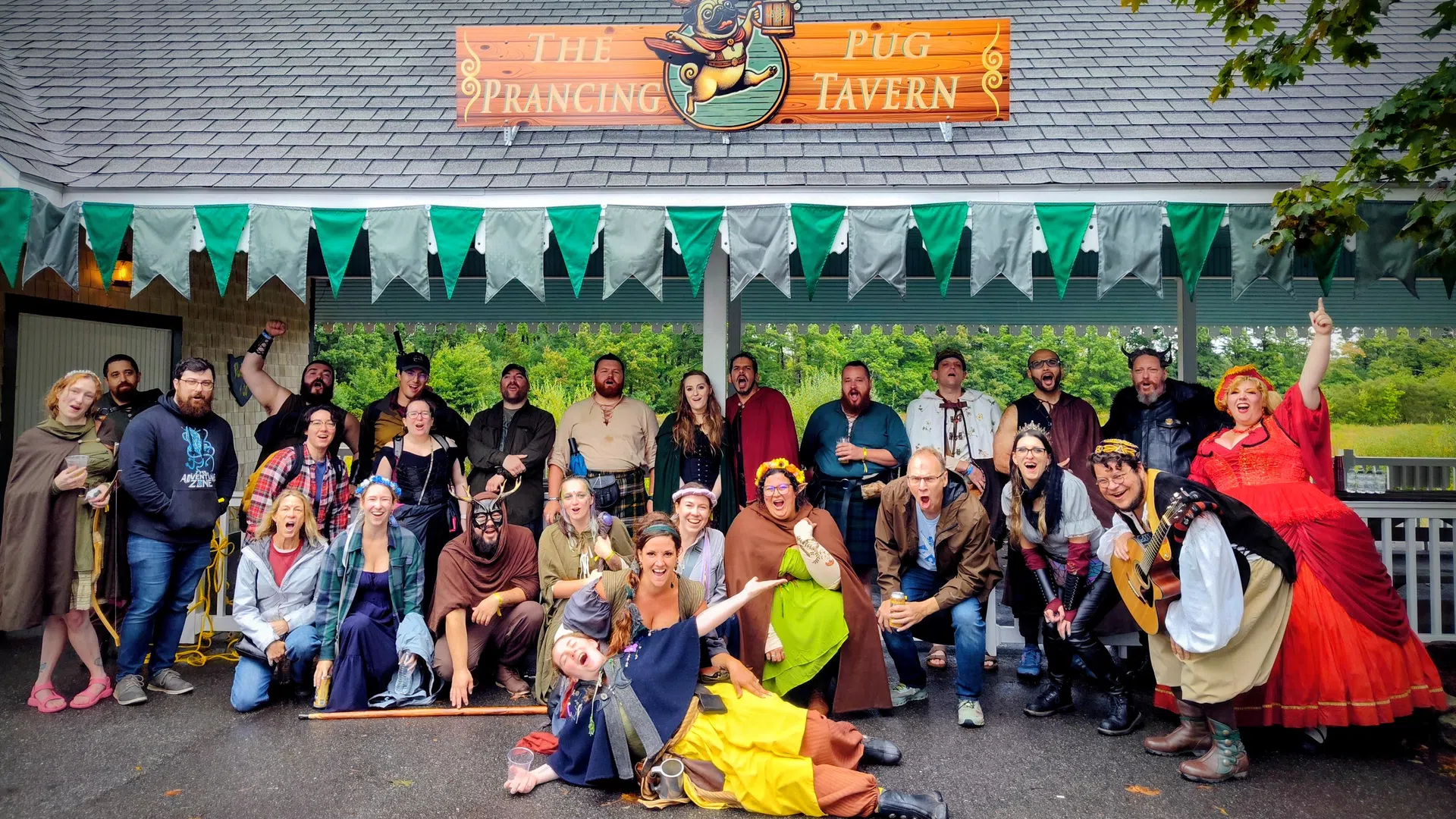 Group of people in colorful medieval costumes posing outside The Prancing Pug Tavern with green bunting overhead.