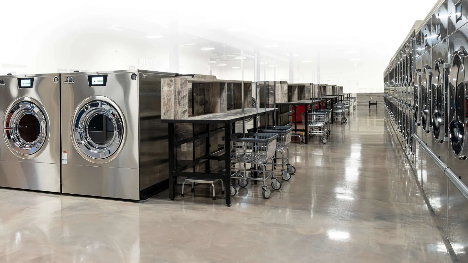 Spacious laundromat with rows of stainless steel washing machines and folding tables with laundry carts.