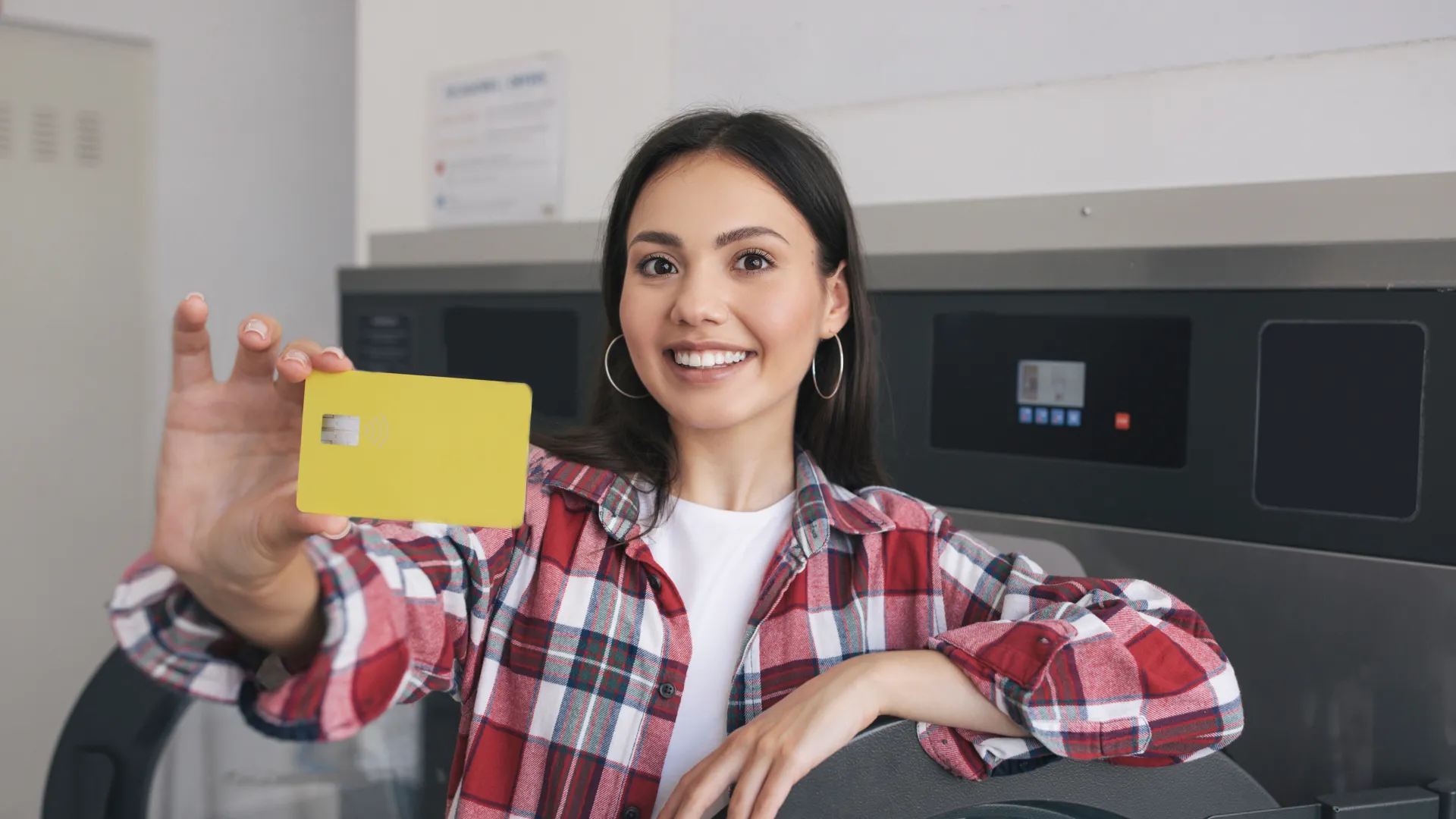 Smiling woman in plaid shirt holding a yellow card at a laundromat with washing machines in the background.