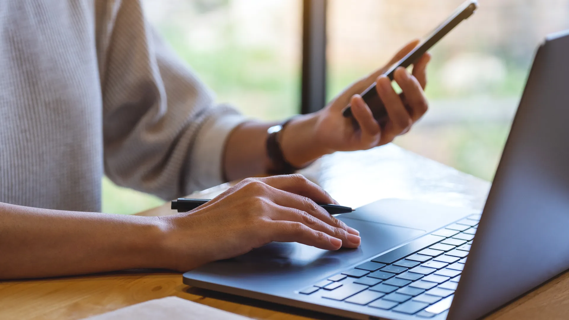 Person using smartphone and laptop simultaneously at wooden desk with natural window light.