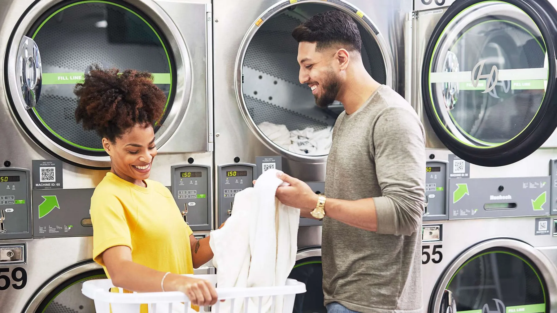Smiling couple doing laundry together at a laundromat with industrial washers and dryers in the background