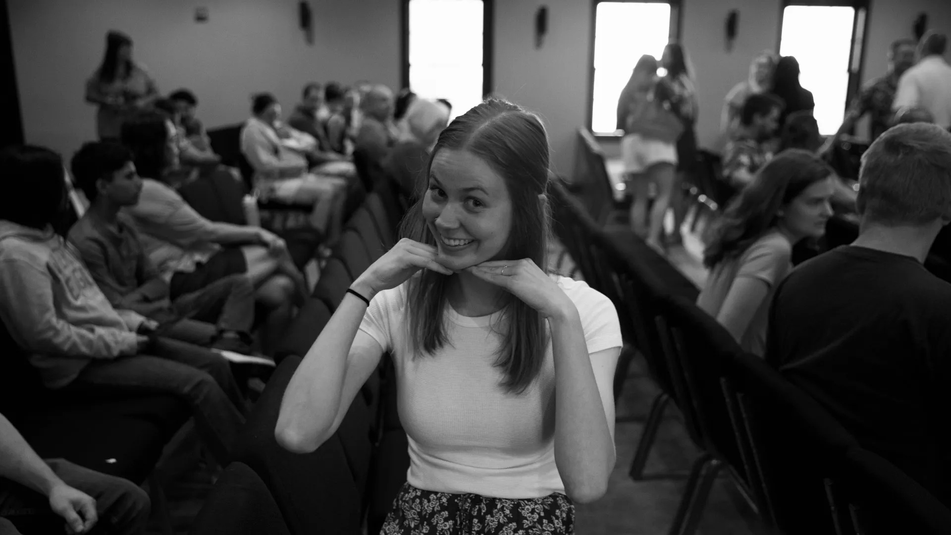 Smiling young woman in white shirt posing with hands under chin in a black and white photo of a room with seated people.