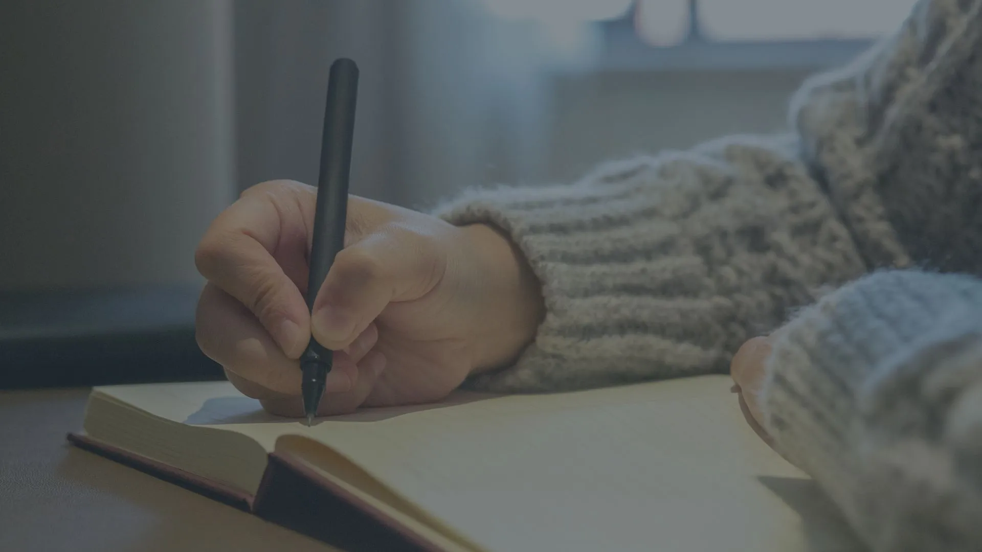 Person wearing a gray knitted sweater writing in an open notebook with a black pen on a table.
