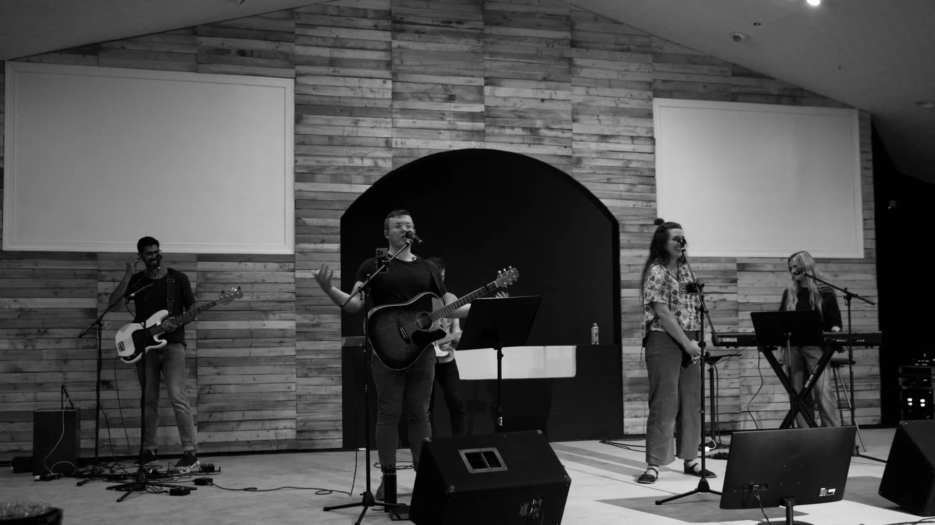 Black and white image of a band performing on stage with guitar, keyboard, and vocalists in a modern wooden-paneled venue.