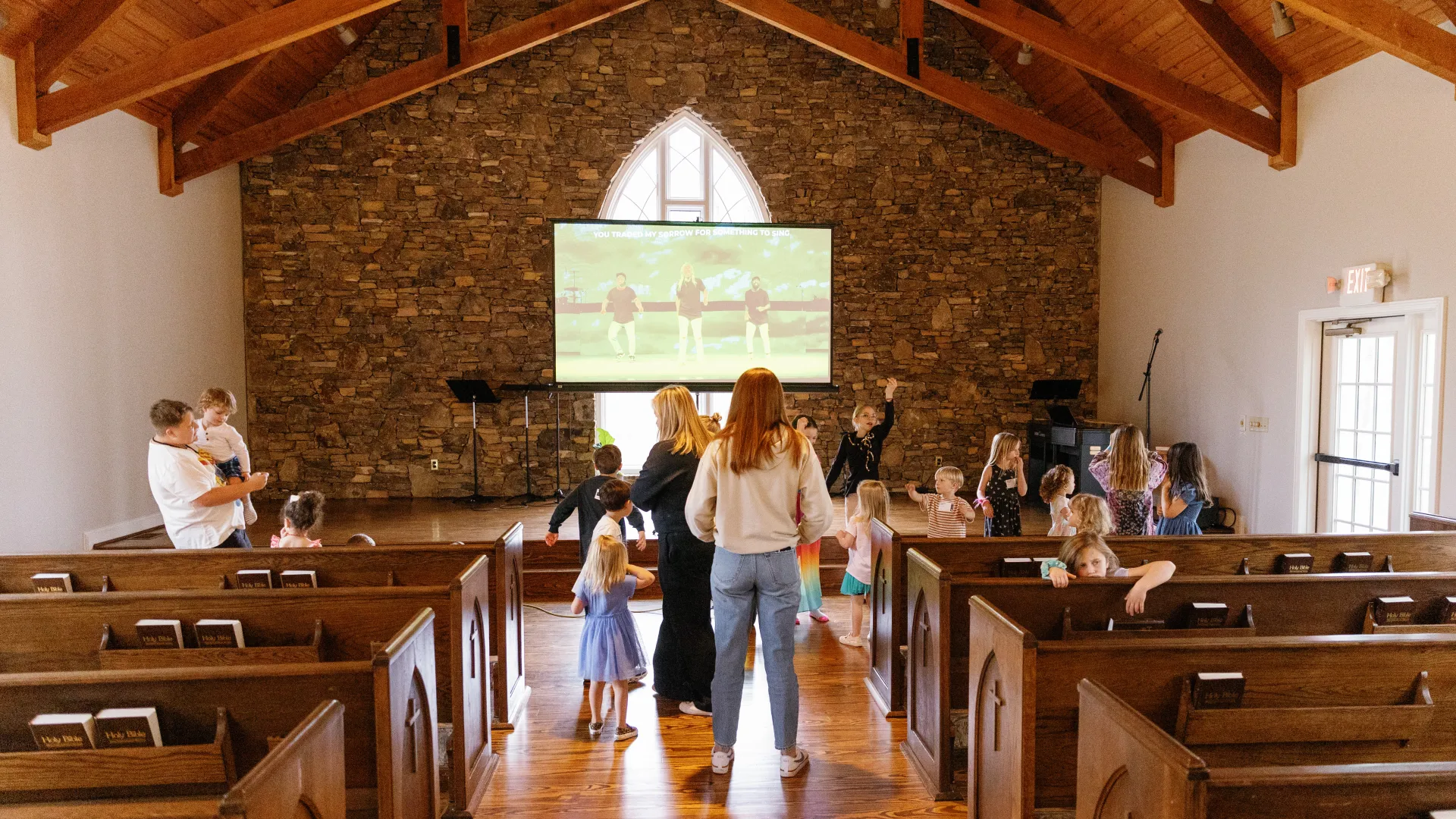 Children and adults watching a screen and interacting inside a wooden church with stone wall and high ceiling.