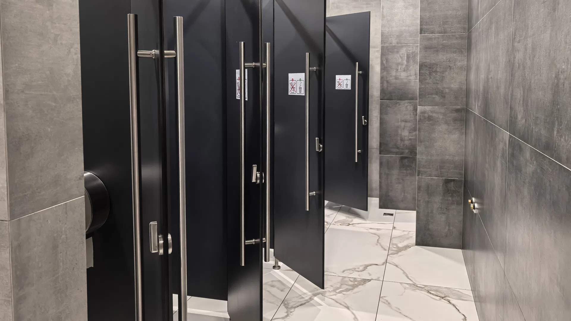 Modern public restroom with multiple black stalls and gray tiled walls and floor.