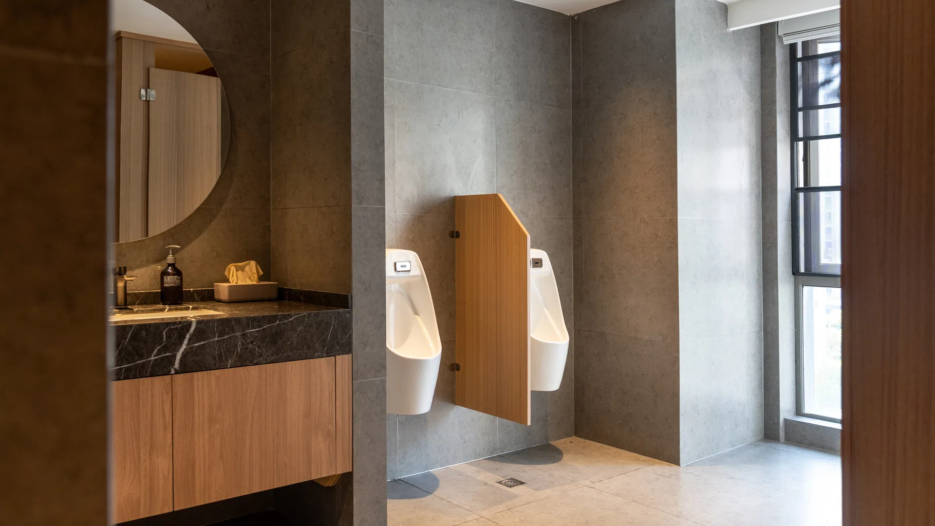 Modern men's restroom with two white urinals and wooden divider next to a sink with mirror and soap dispenser.