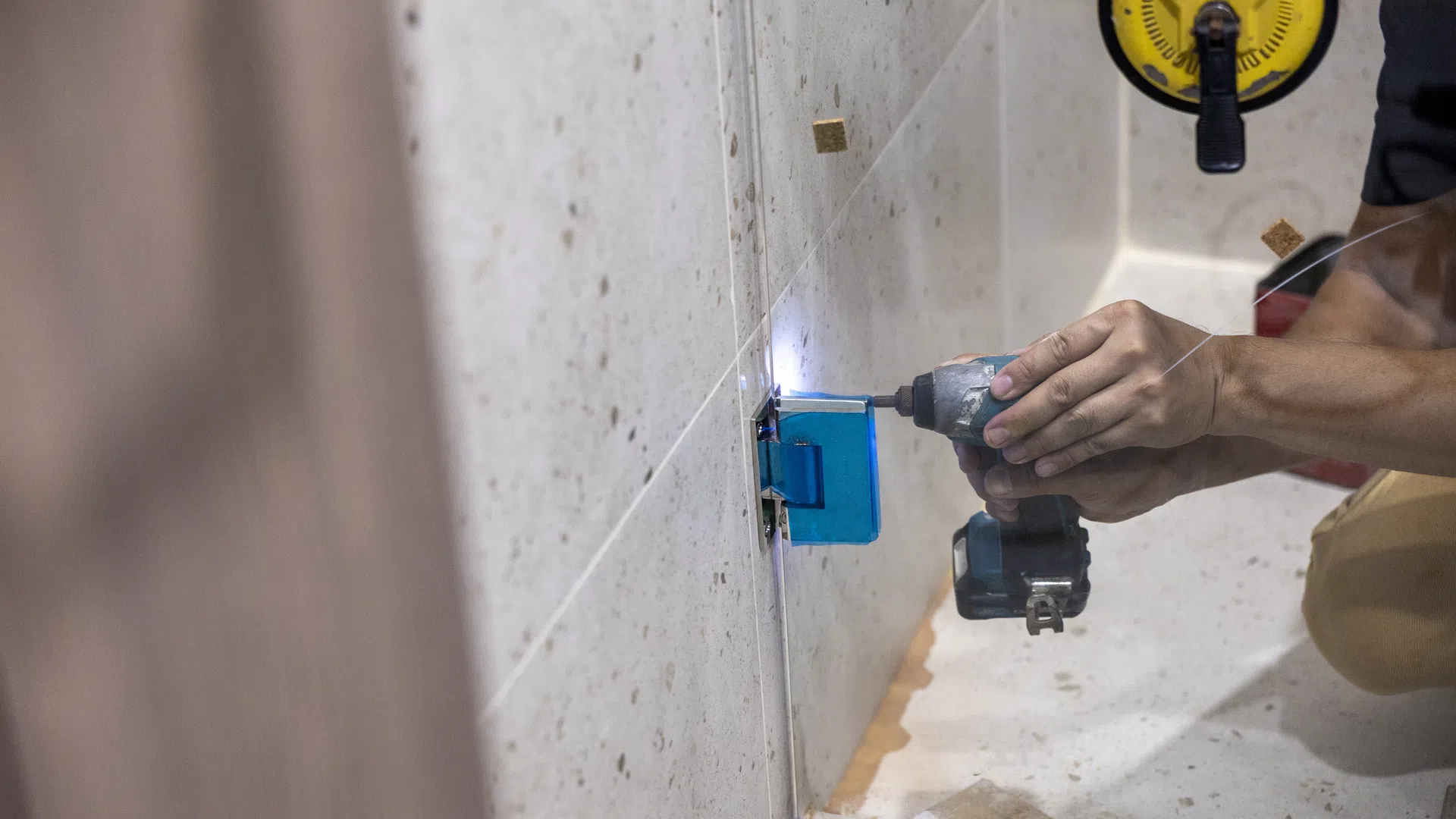 Person using a cordless drill to install a glass shower door in a bathroom under construction.