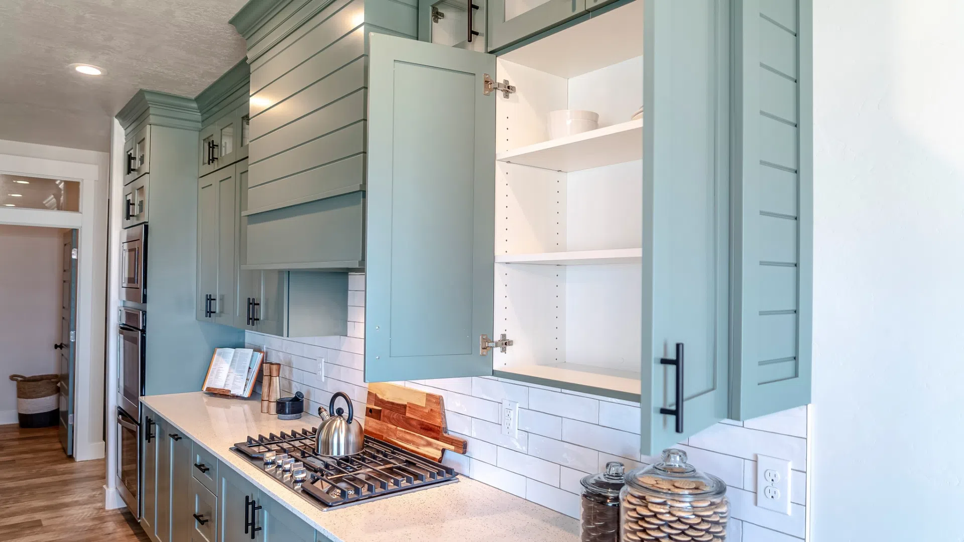 Modern kitchen with teal cabinets, white subway tile backsplash, gas stove, and countertop jars under bright lighting.
