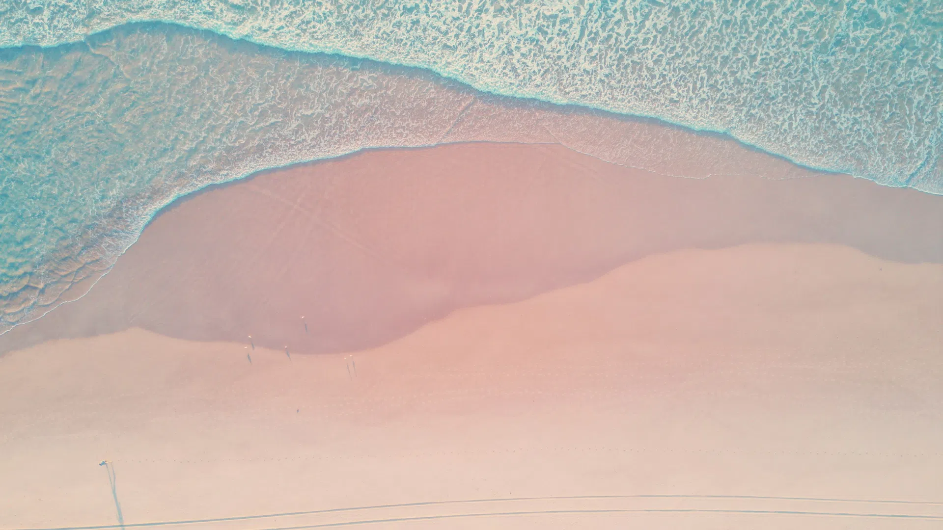 Aerial view of turquoise ocean waves gently washing onto a wide pink sandy beach with faint shadows and tire tracks.