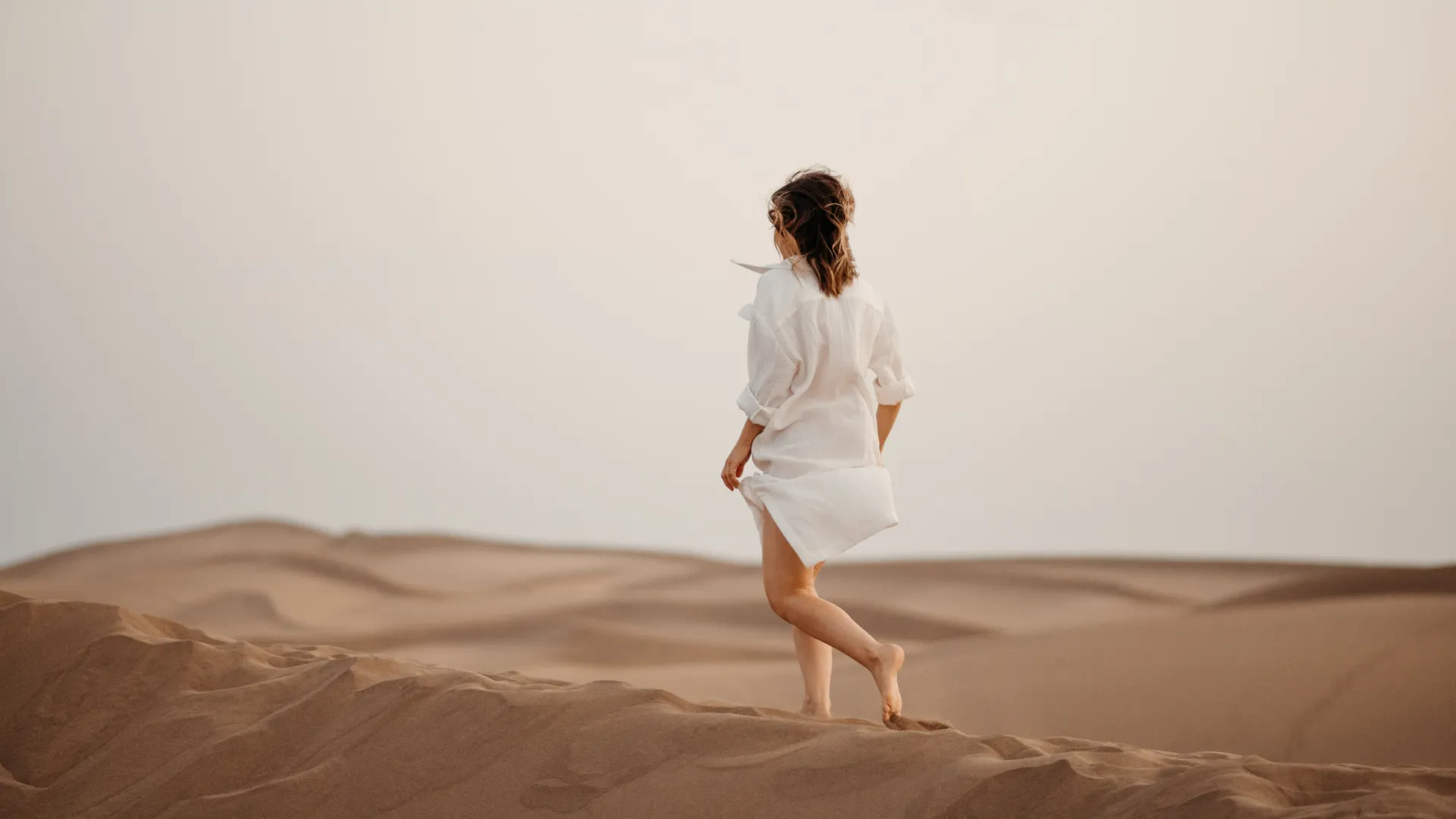 Woman in white shirt walking barefoot across sandy desert dunes at sunset with soft lighting.