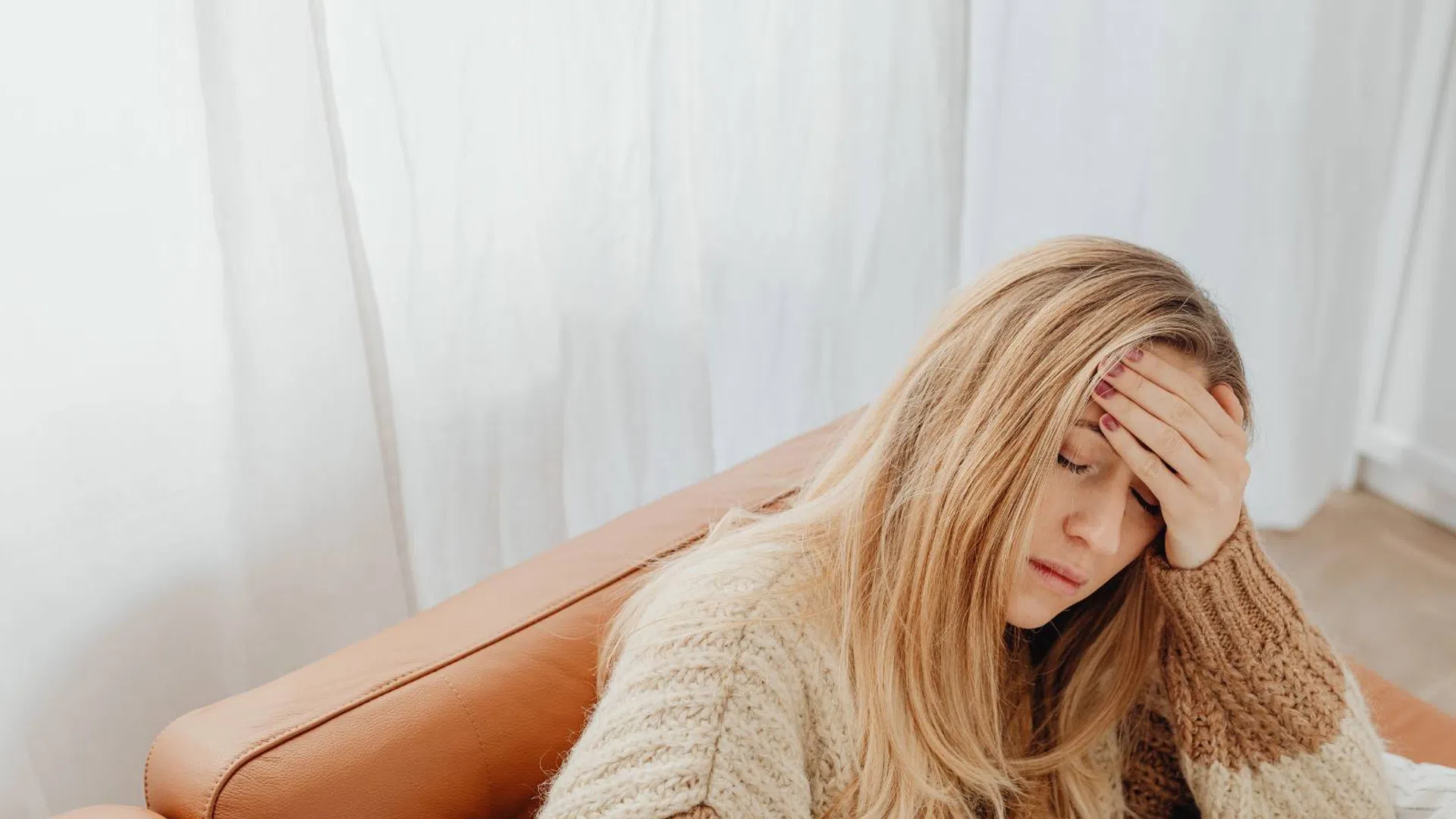 Stressed woman with blonde hair in a cozy sweater sitting on a brown couch holding her forehead.