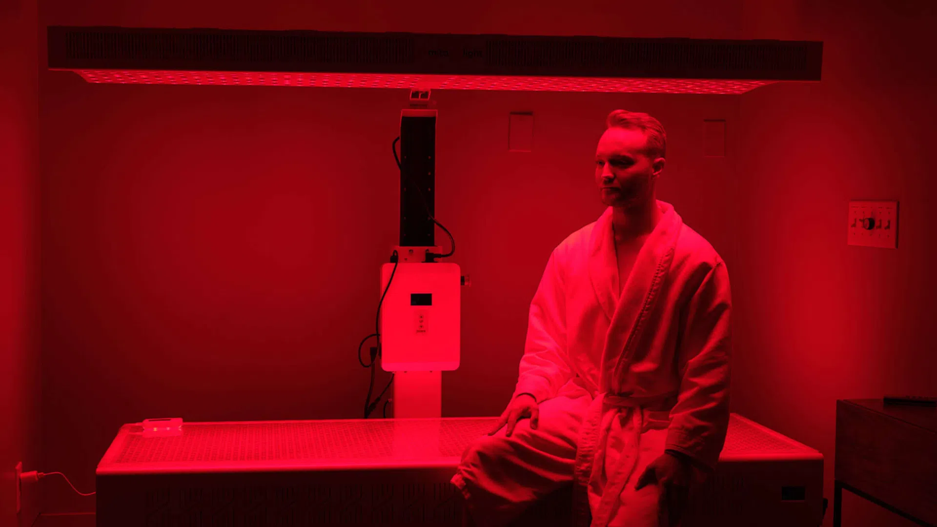 Man in white robe sitting on medical therapy bed under red light in clinical room.