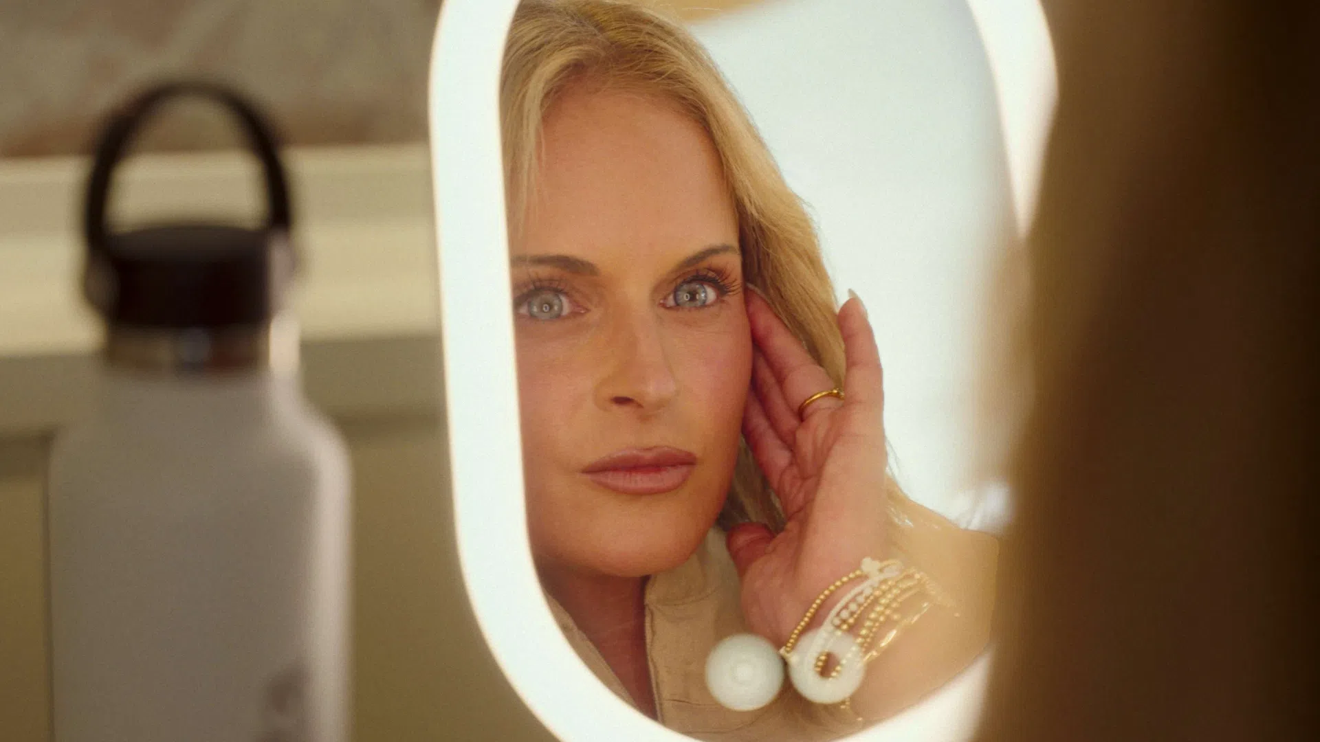 Blonde woman with blue eyes looking at her reflection in a lighted vanity mirror, wearing gold rings and bracelets.