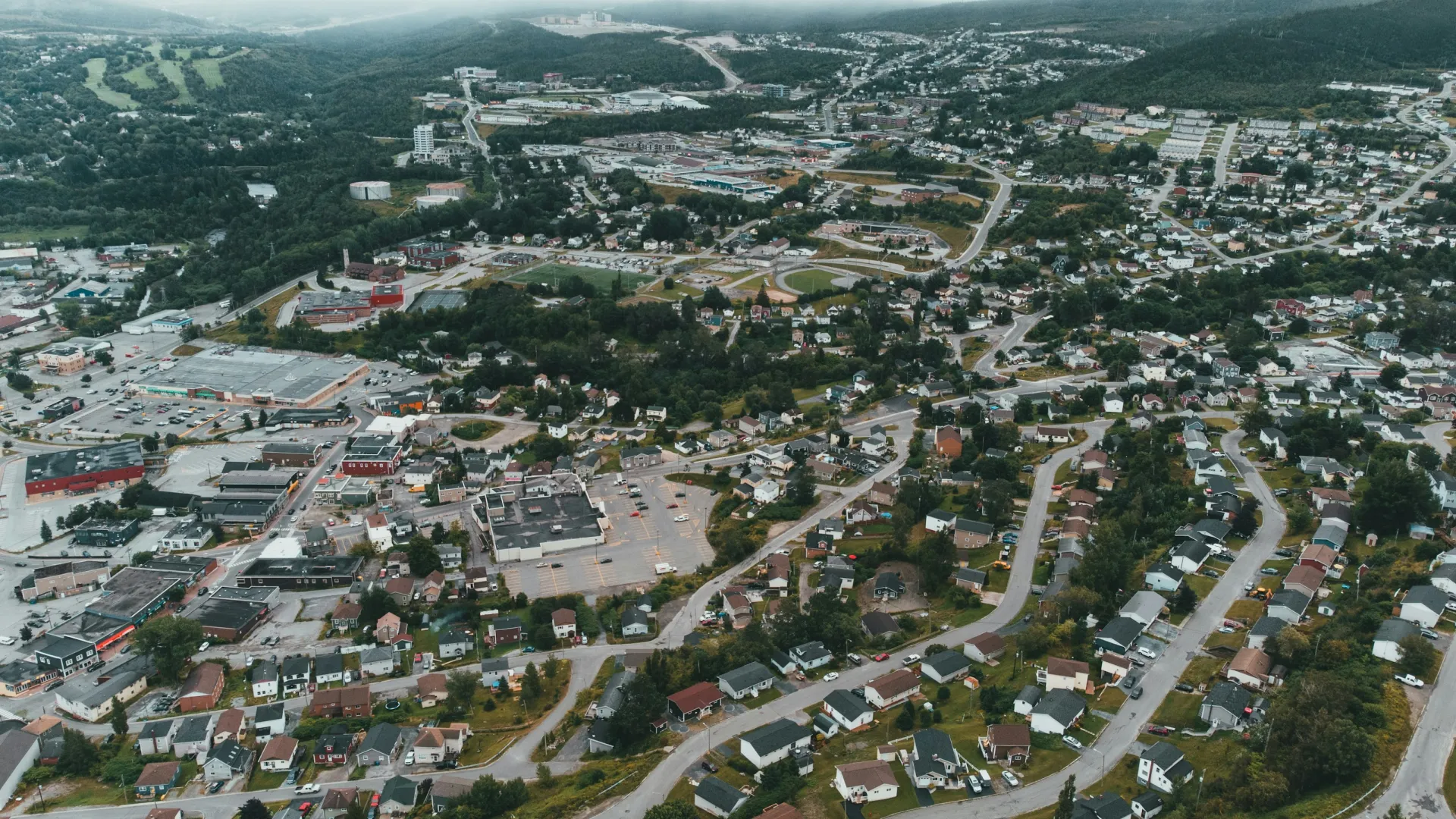 Aerial view of a suburban neighborhood with houses, roads, commercial areas, and surrounding green hills under a cloudy sky.
