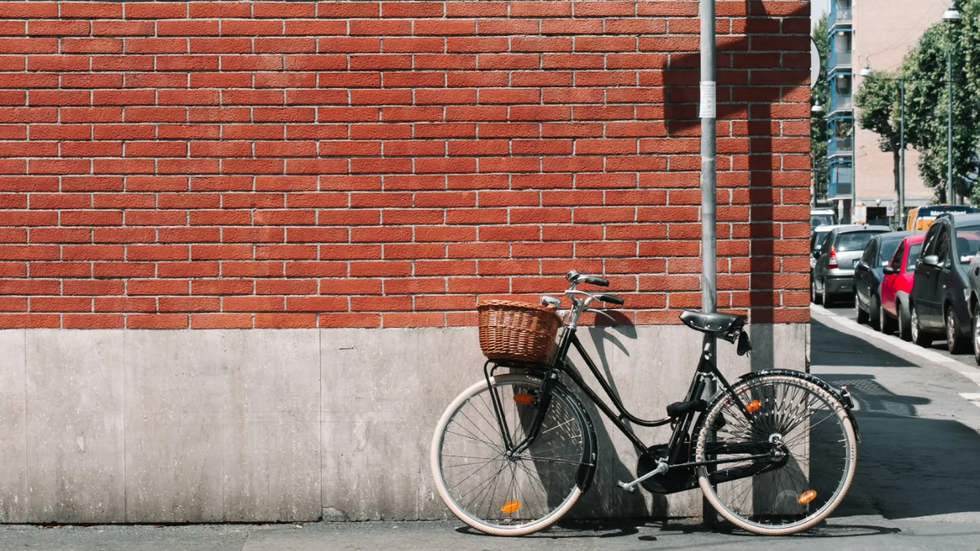 Black bicycle with a basket parked against a red brick wall near a graffiti-covered street sign on a sunny day.