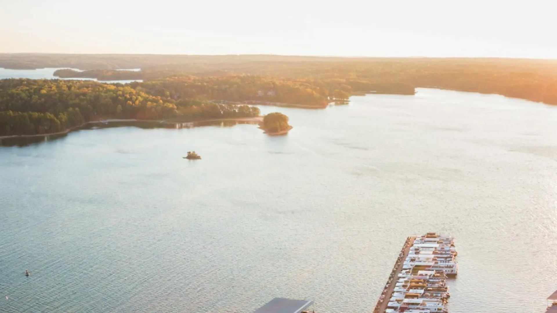 Aerial view of a marina with docks, boats, and calm water during sunset surrounded by autumn trees.