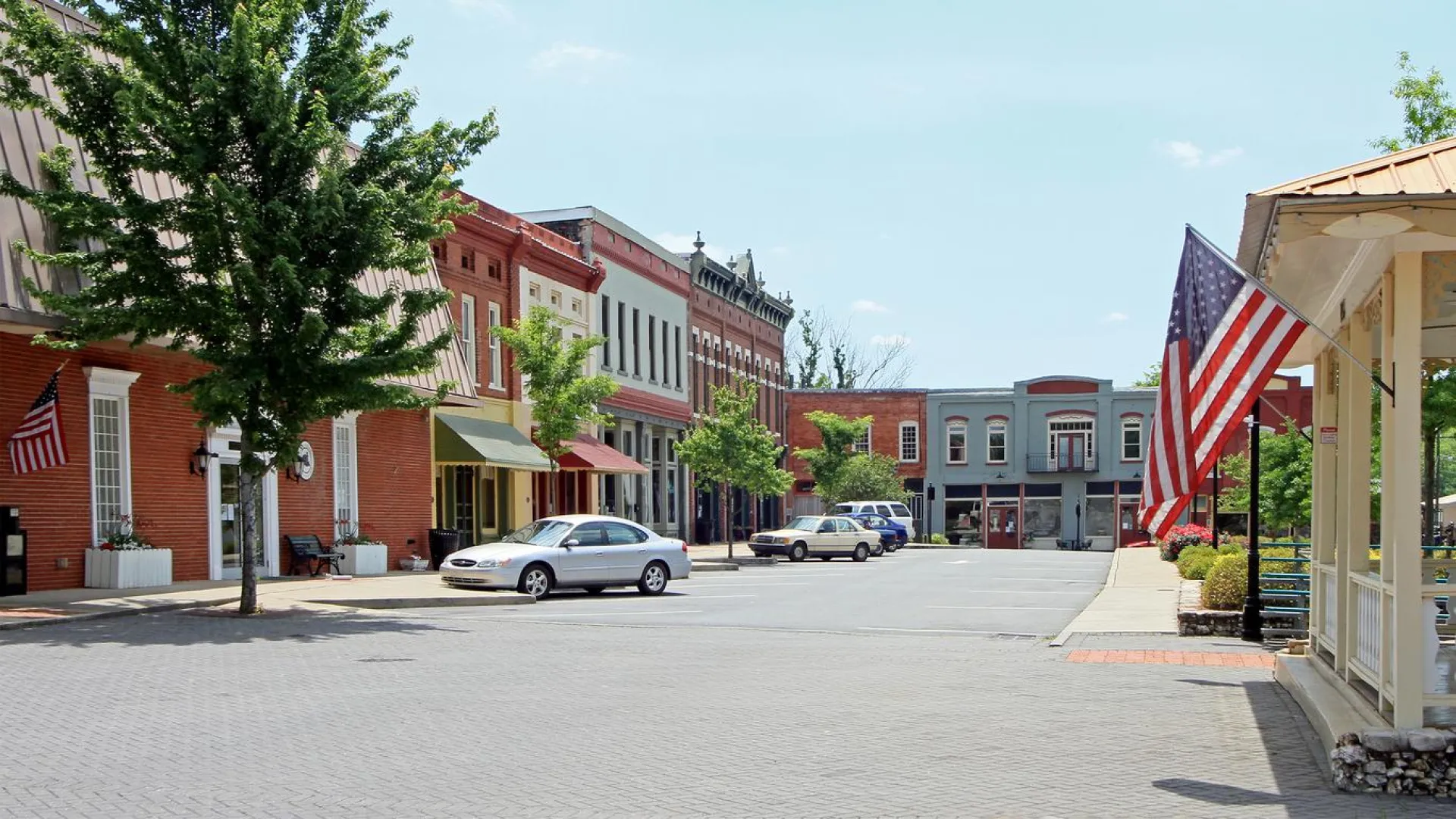 Small town main street with historic brick buildings, parked cars, trees, and American flags on a sunny day.